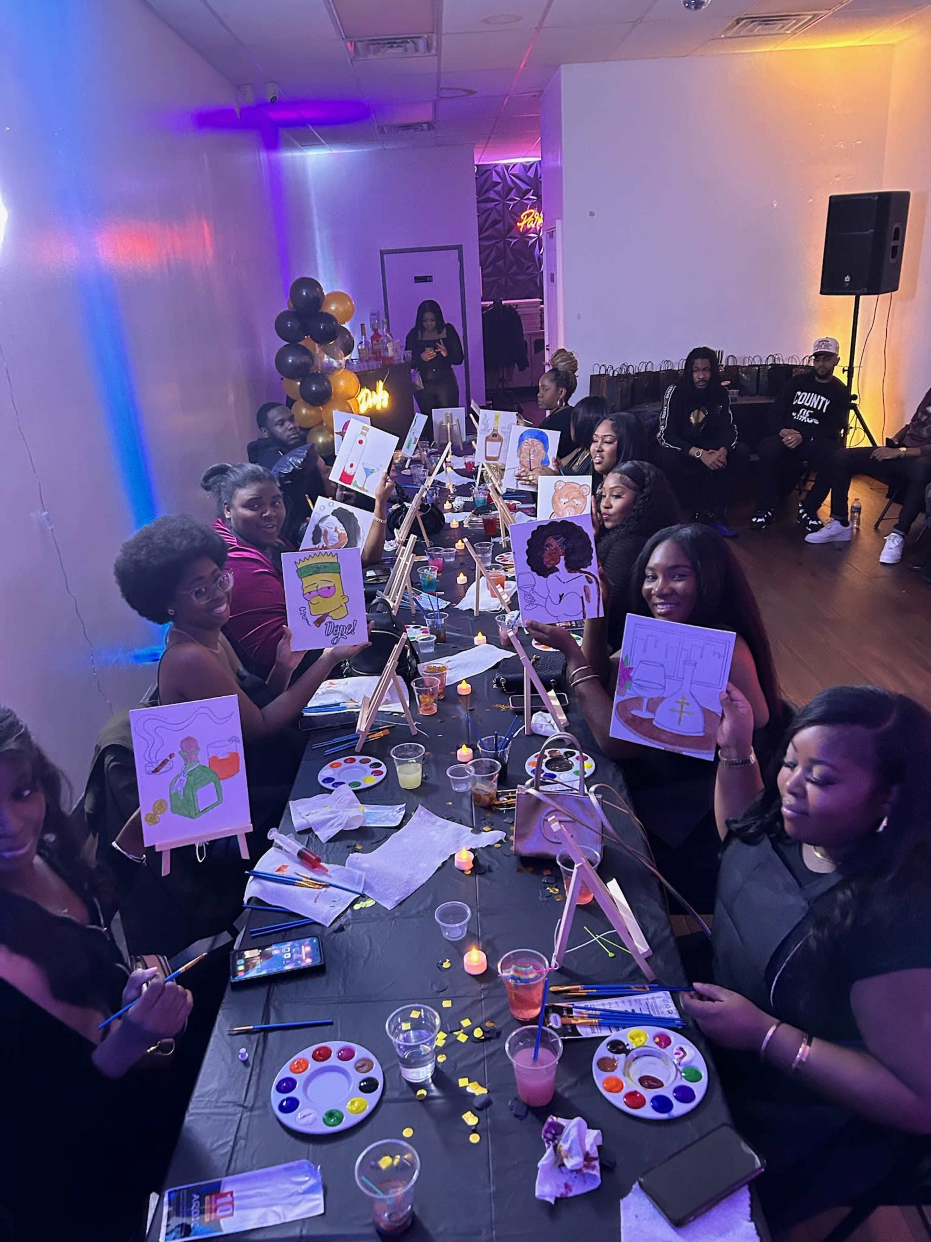 A group of women are seated at tables in a brightly lit room, holding up their paintings while surrounded by art supplies and party decorations.