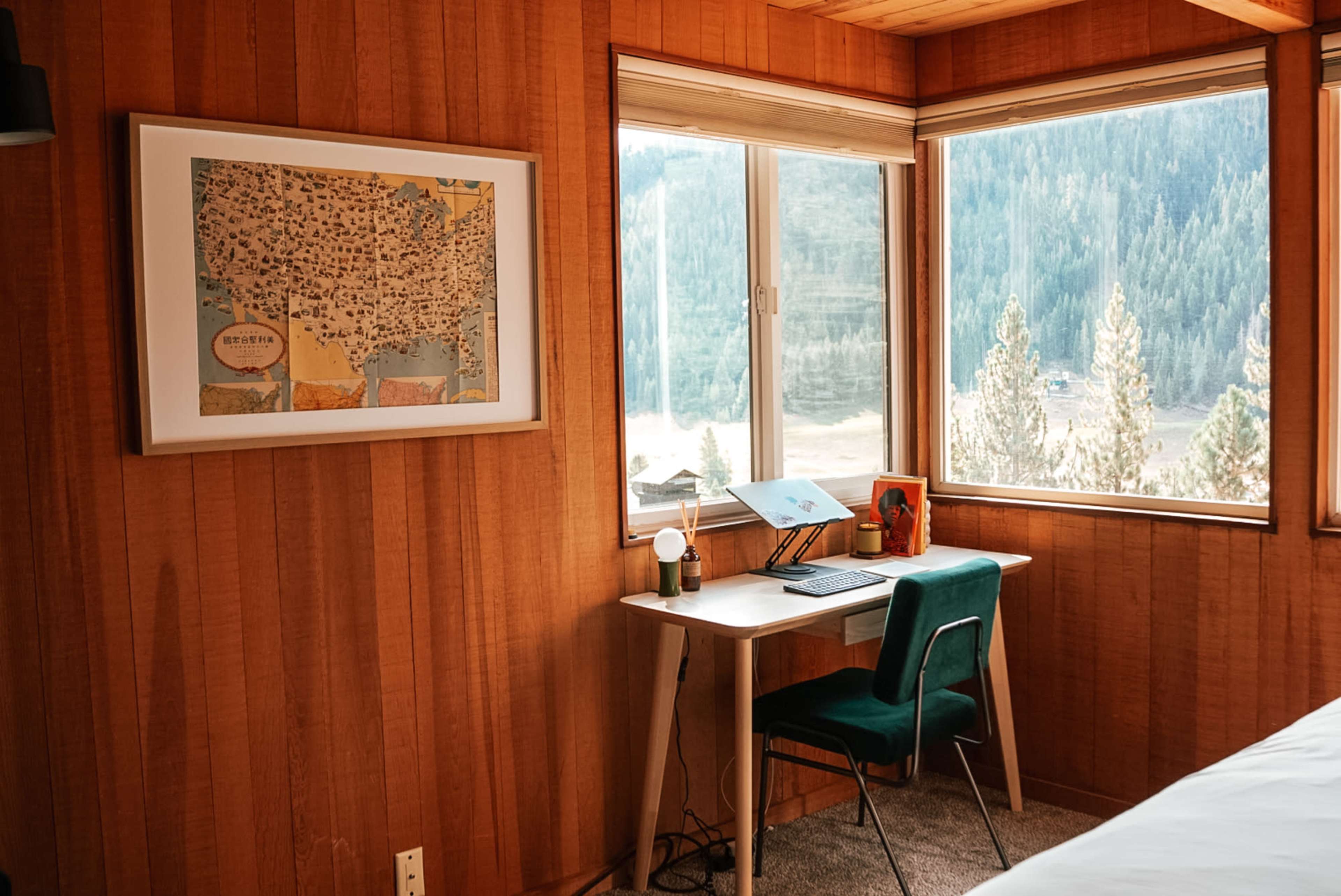 The image shows a wooden-paneled room with a desk, chair, and a map framed on the wall, overlooking a scenic view of trees and mountains through large windows.