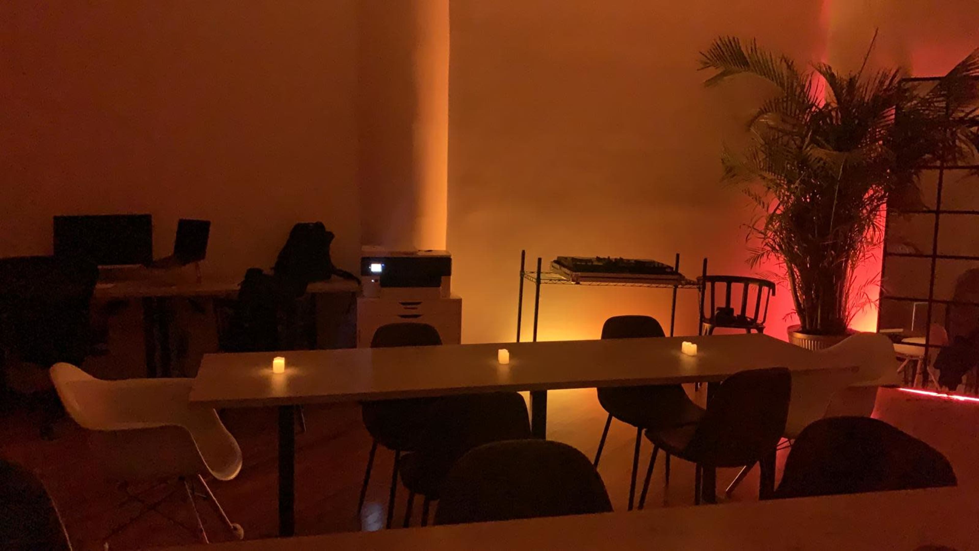 A dimly lit room features a long table surrounded by chairs, with a printer and music equipment in the background and soft lights illuminating a nearby plant.