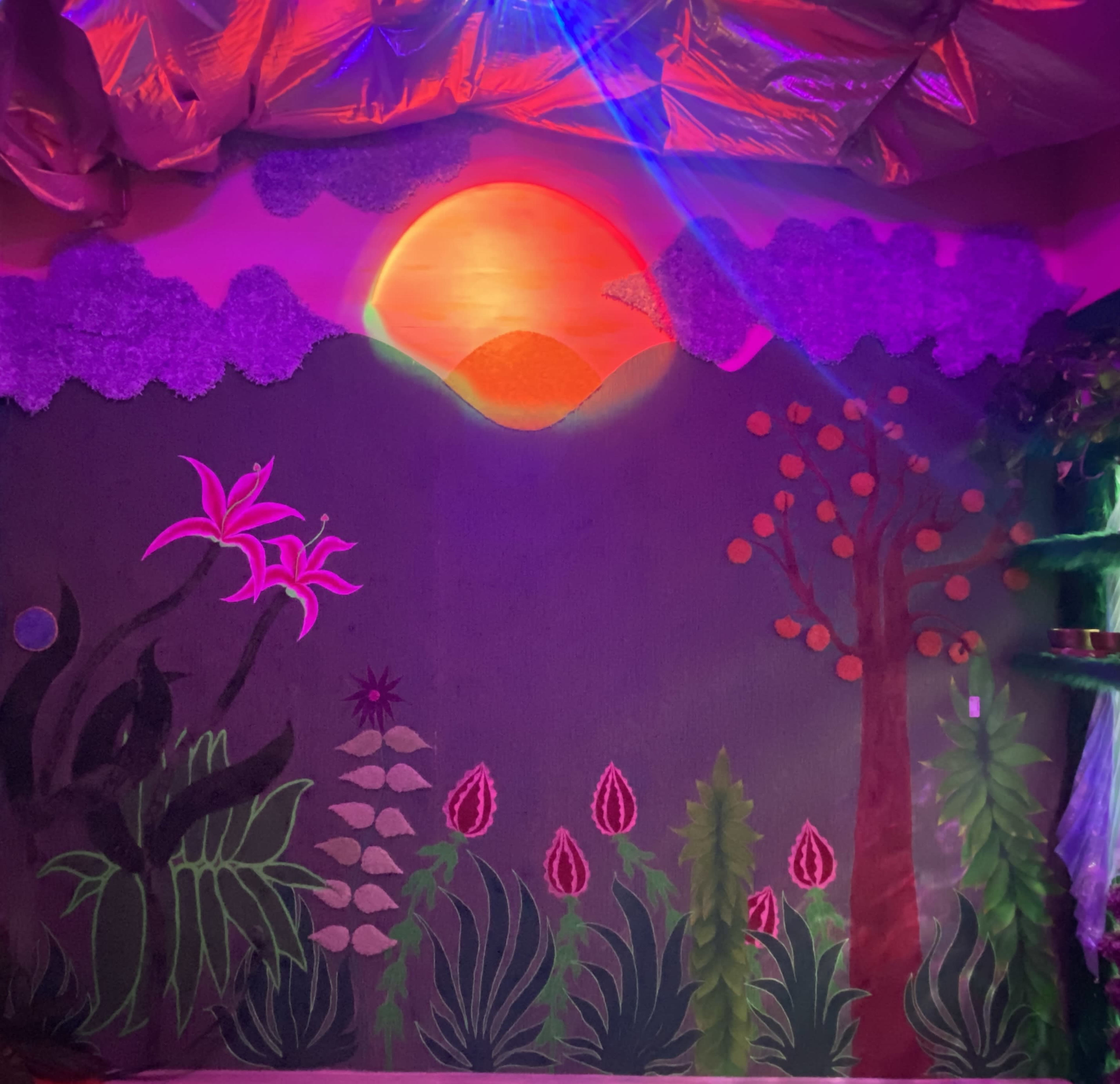 The image features a colorful mural with a sunset, stylized plants, and flowers on a wall, illuminated by vibrant lighting.