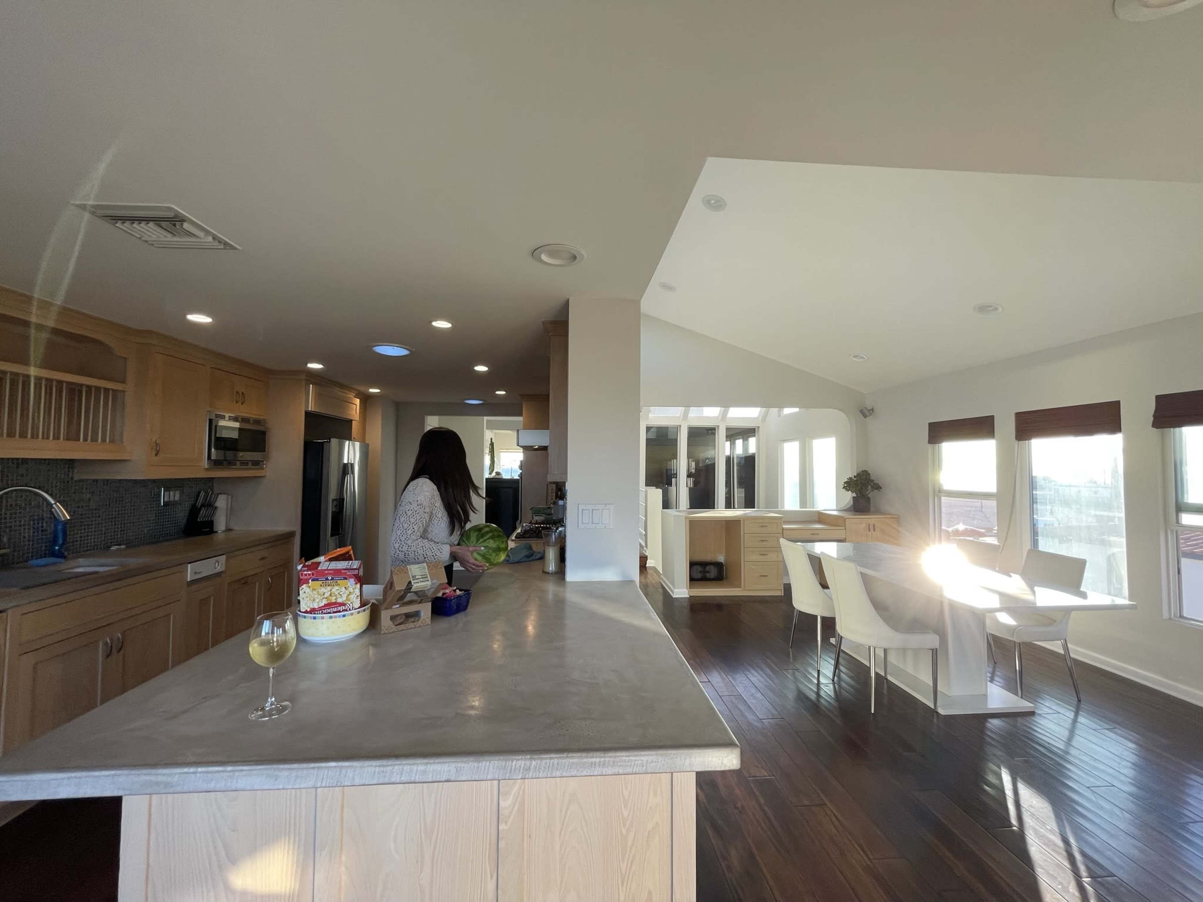 A woman stands at a kitchen counter with various items while sunlight streams through large windows in an open-concept living space.
