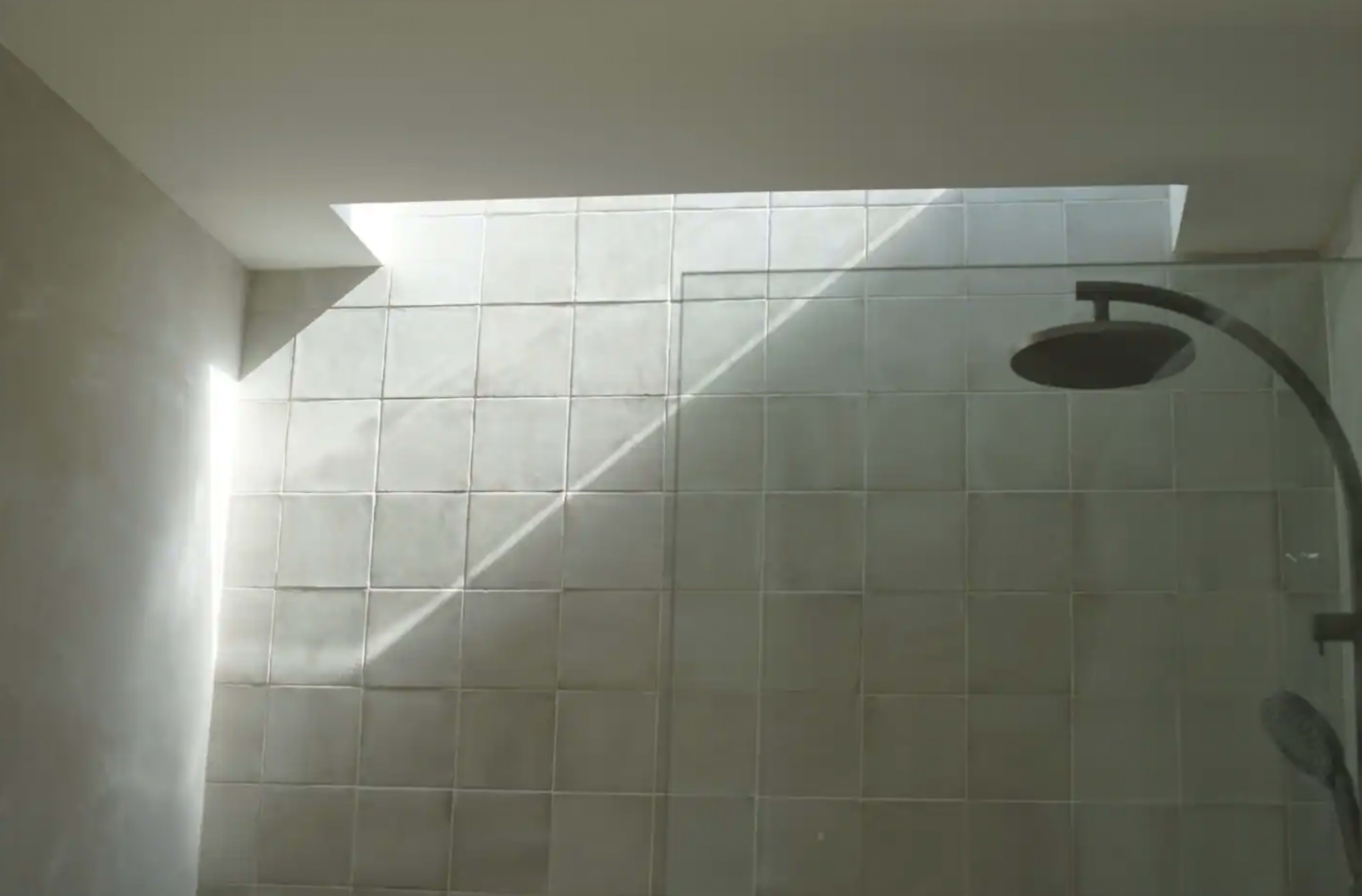 A shower space features tiled walls with natural light streaming from a skylight, creating a diagonal shadow across the surface.