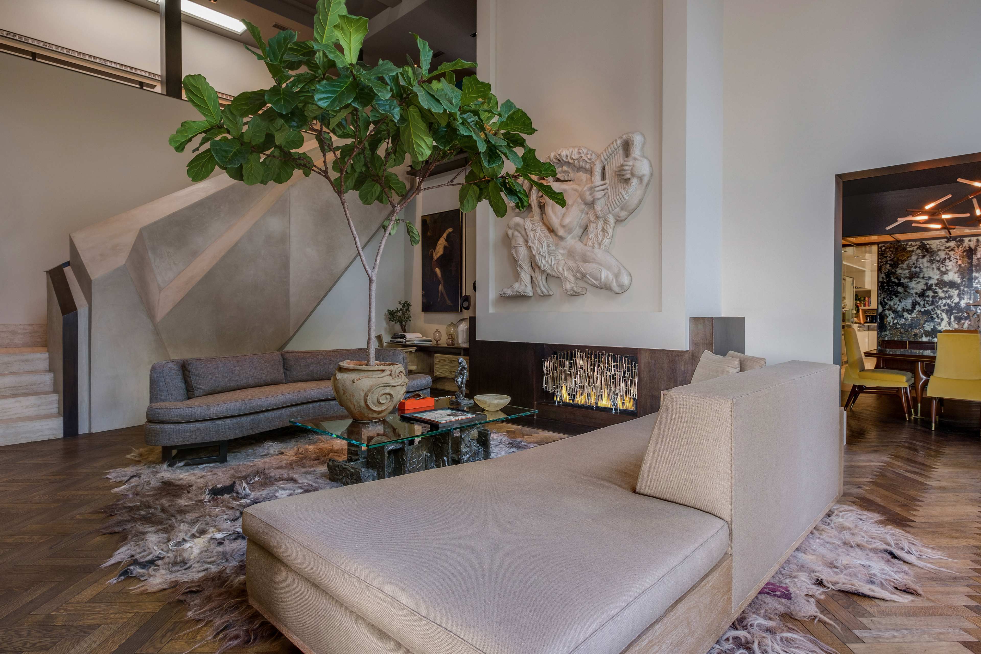 The image shows a modern living room with a large, light-colored sectional sofa, a decorative indoor plant, and a sculpture on the wall, alongside a coffee table and a rug on a wooden floor.