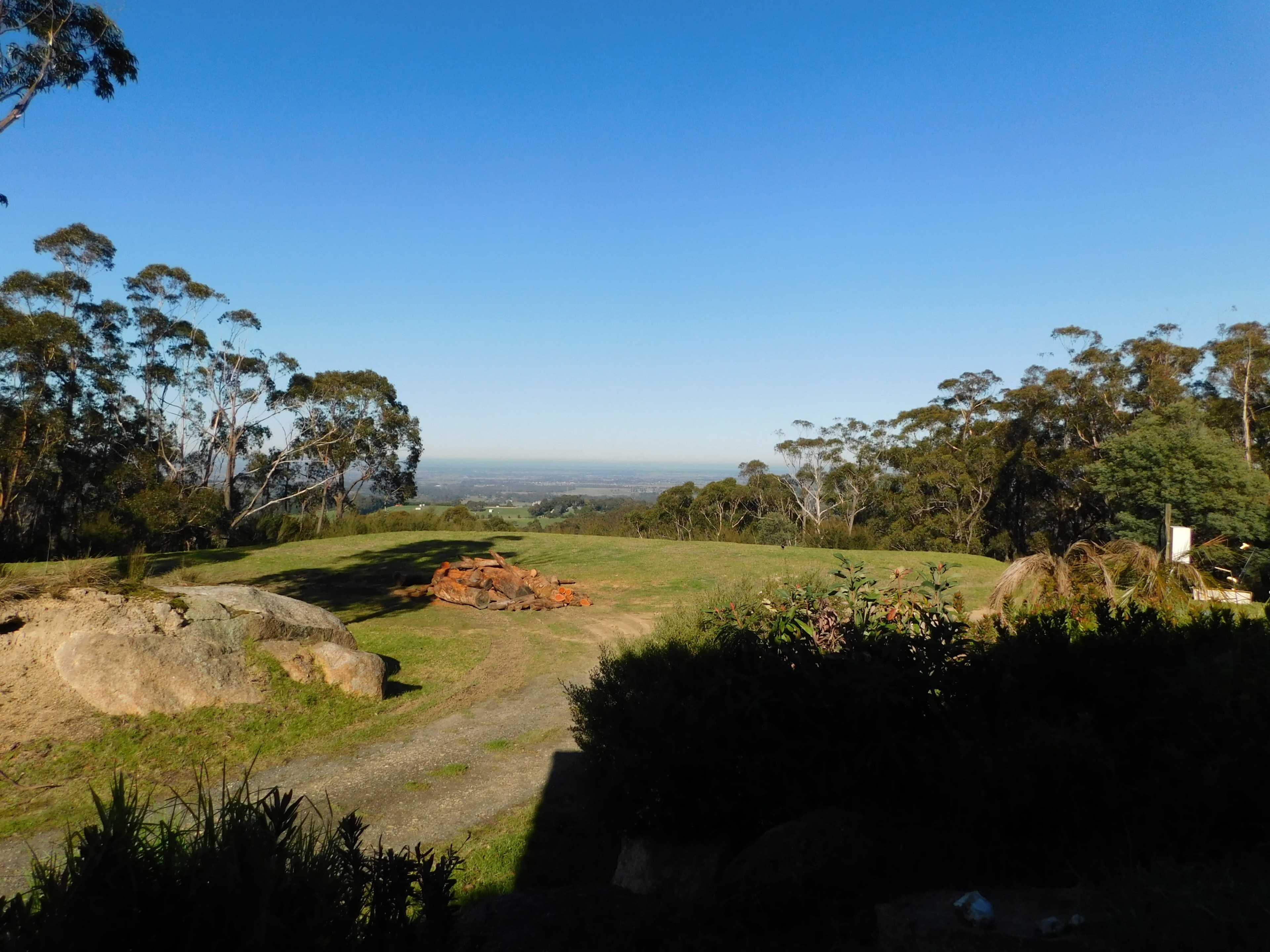 A grassy landscape with large rocks and trees, overlooking a distant view under a clear blue sky.