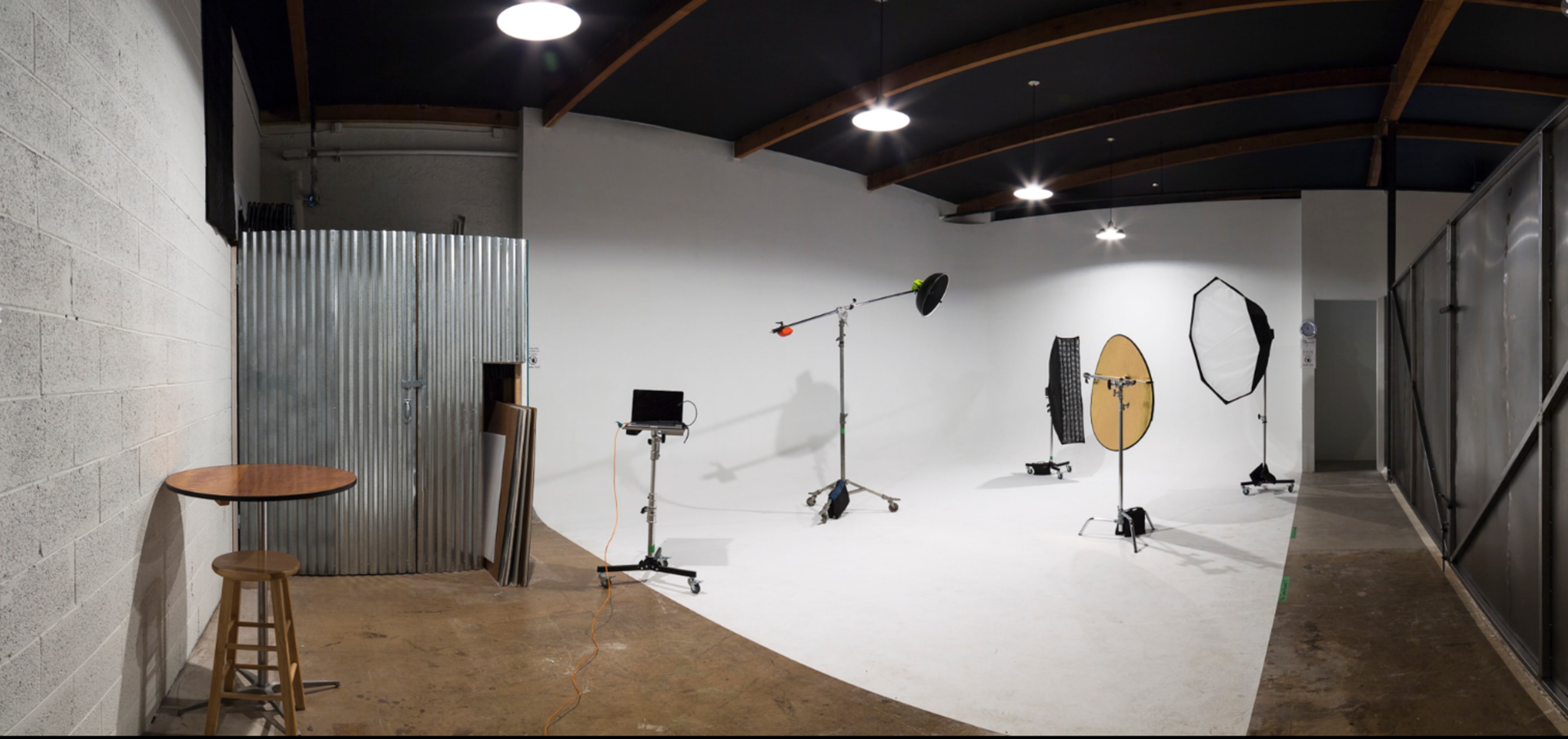 The image shows a photography studio with a seamless white backdrop, several lighting equipment setups, and a small bar table in the corner.