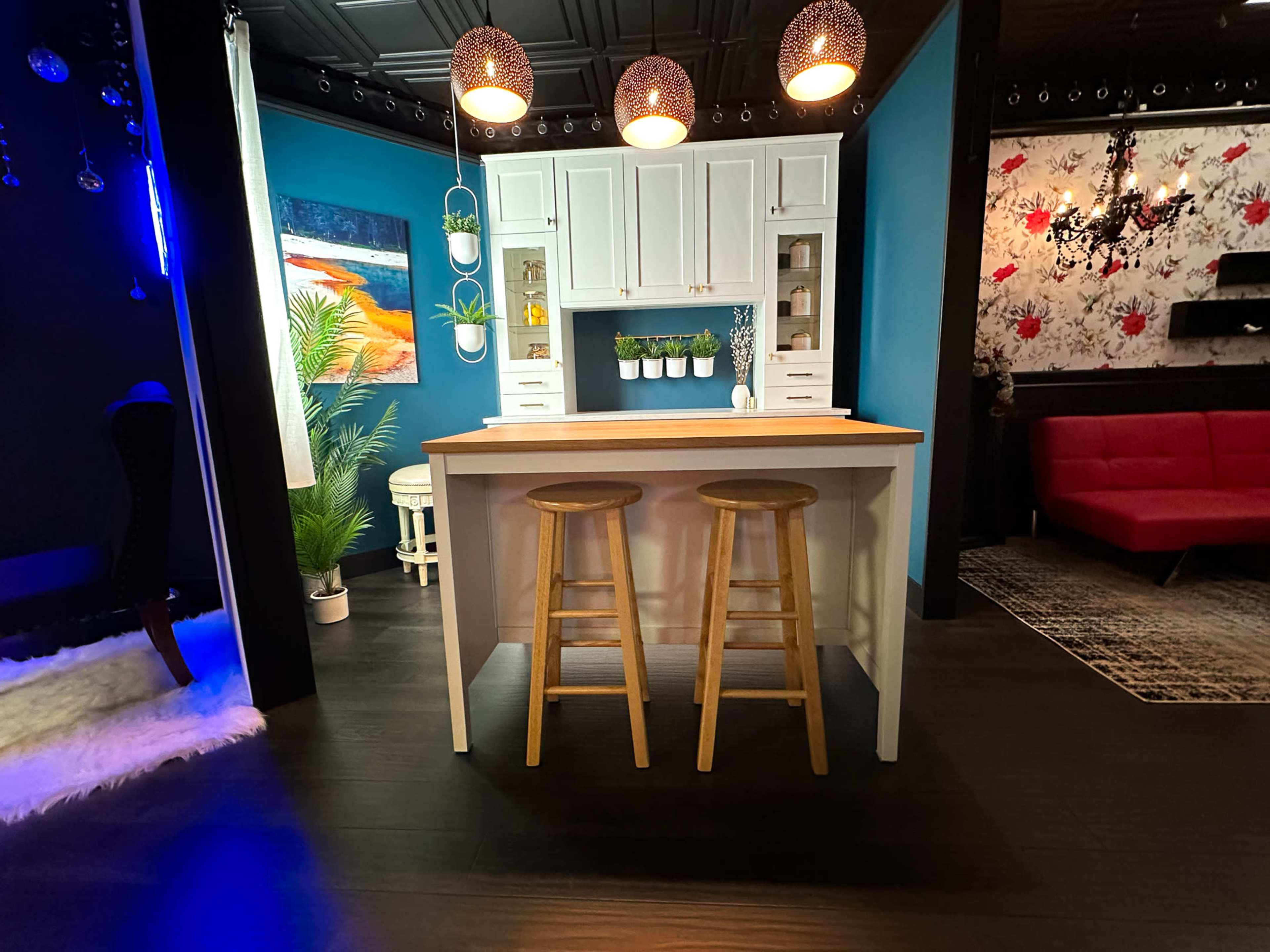 The image shows a modern kitchen space with a white island and two wooden stools, surrounded by vibrant blue and black walls, decorative lighting, and various ornamental details.
