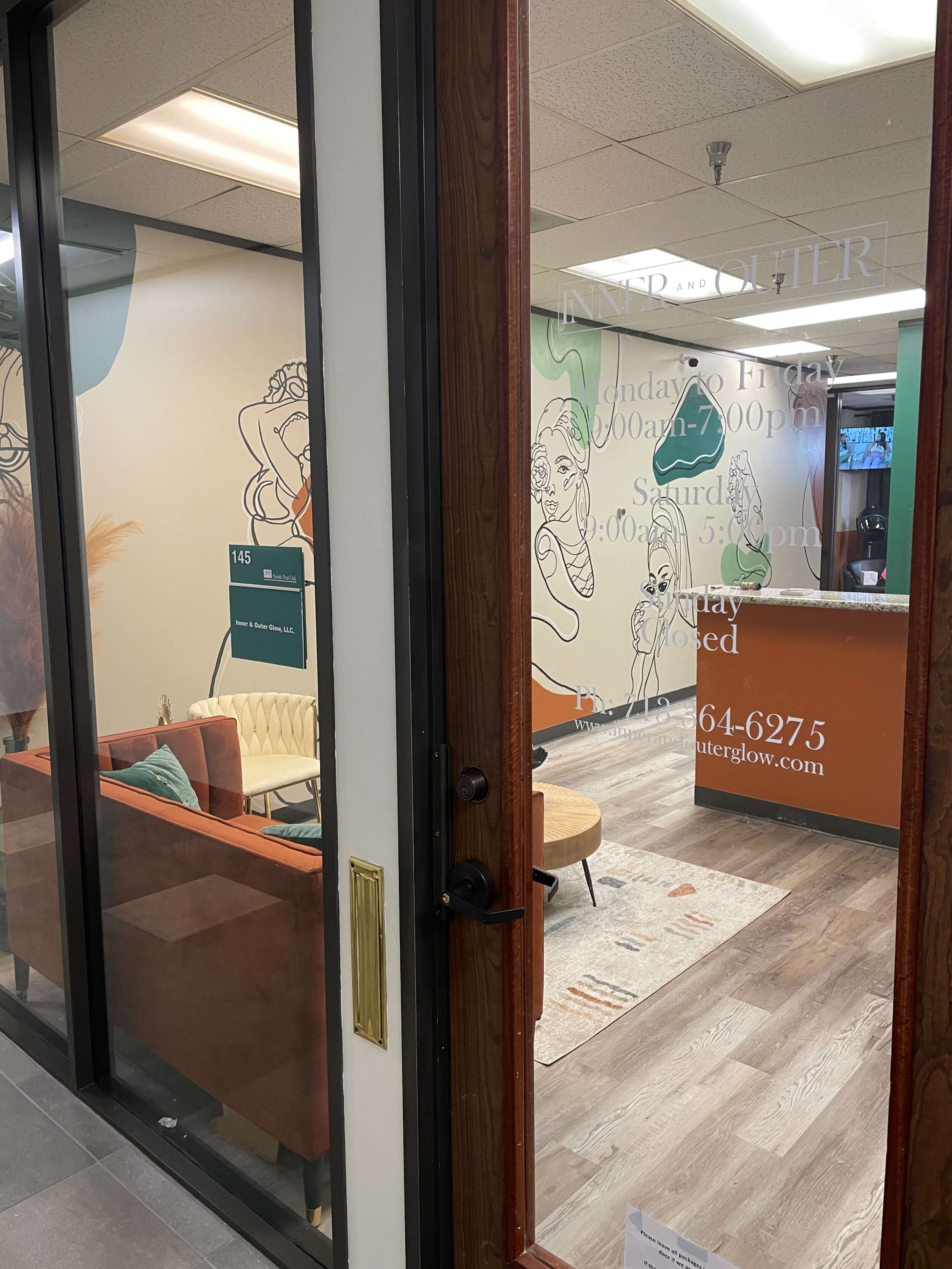 An entrance to a modern office with a welcoming reception area visible through a glass door, featuring a sofa and decorative artwork on the walls.