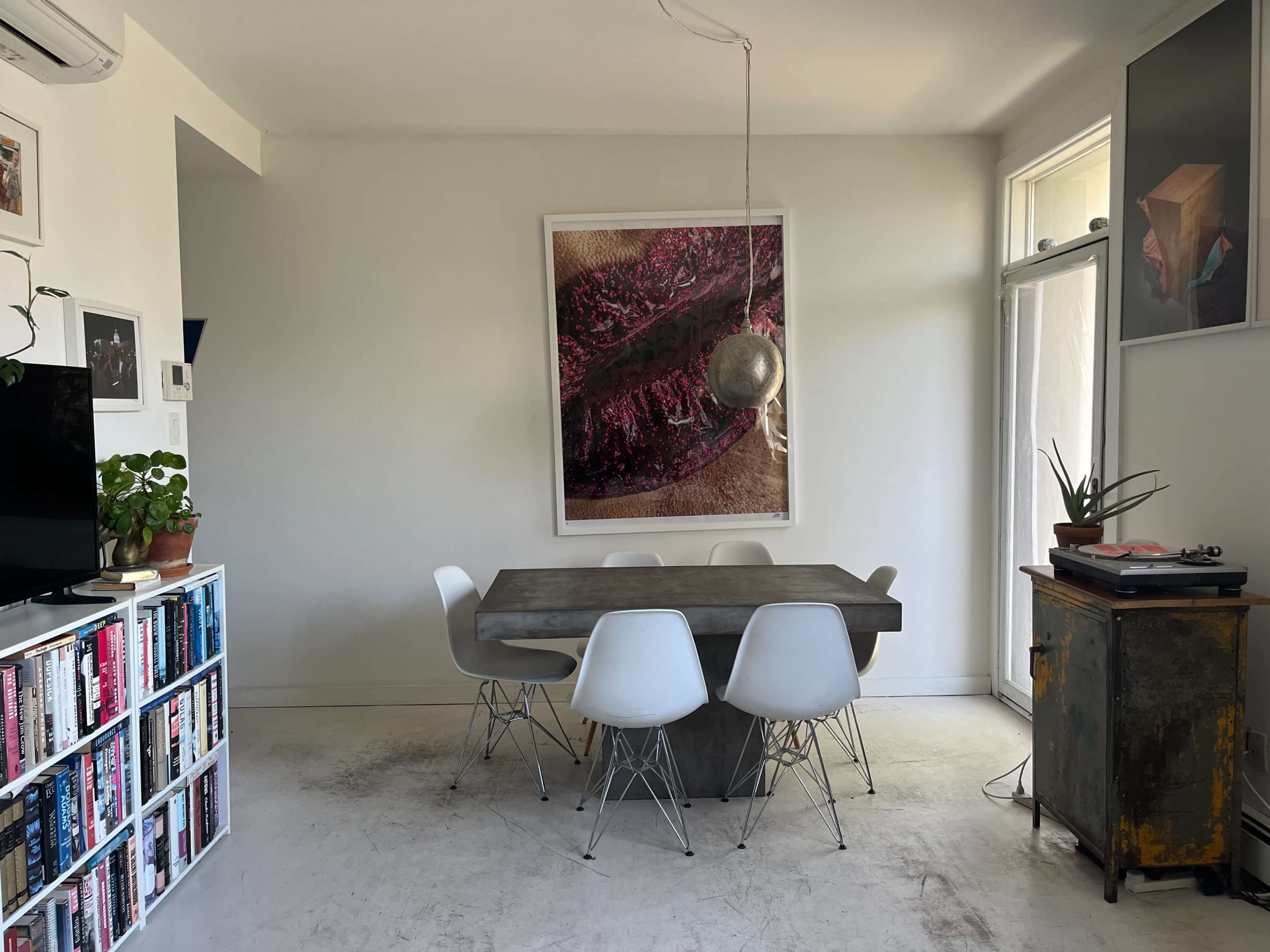 The image shows a minimalist dining area with a large art piece on the wall, a concrete dining table surrounded by white chairs, and a bookshelf filled with books.