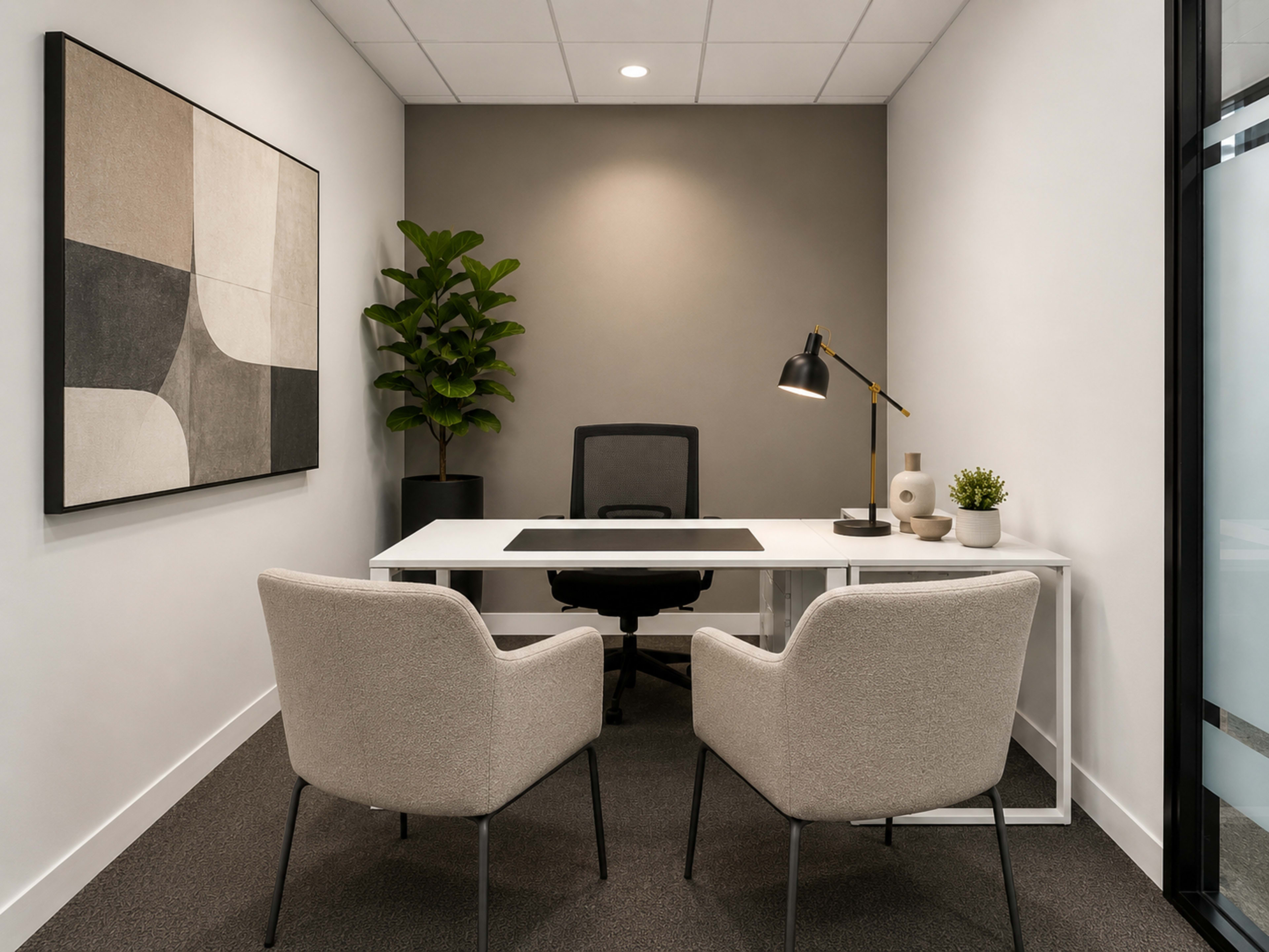 The image shows a modern office space with a white desk, two light-colored chairs, a small plant, and a framed abstract artwork on the wall.