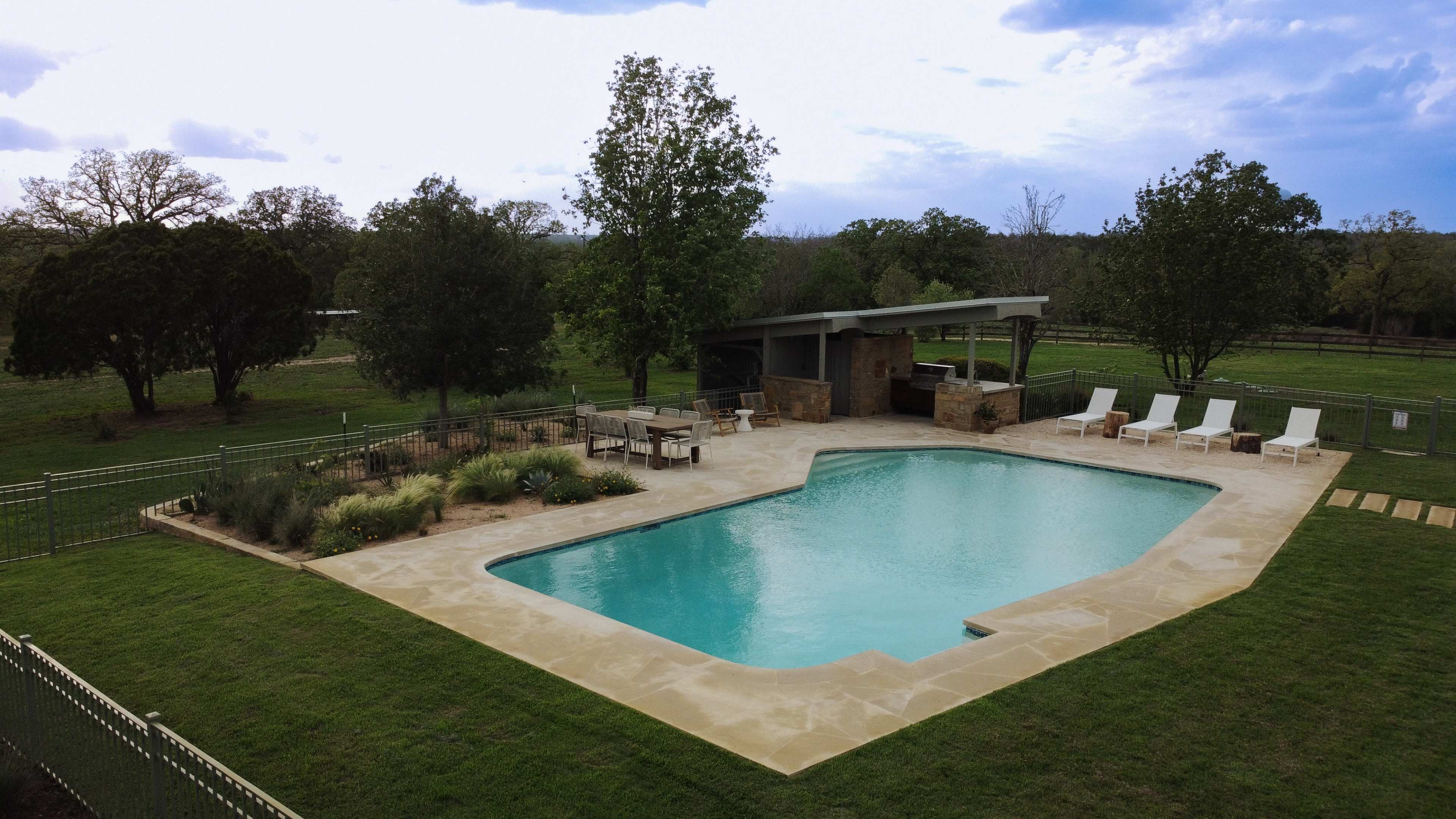 A rectangular swimming pool with a wooden gazebo and lounge chairs is situated in a grassy area surrounded by trees.