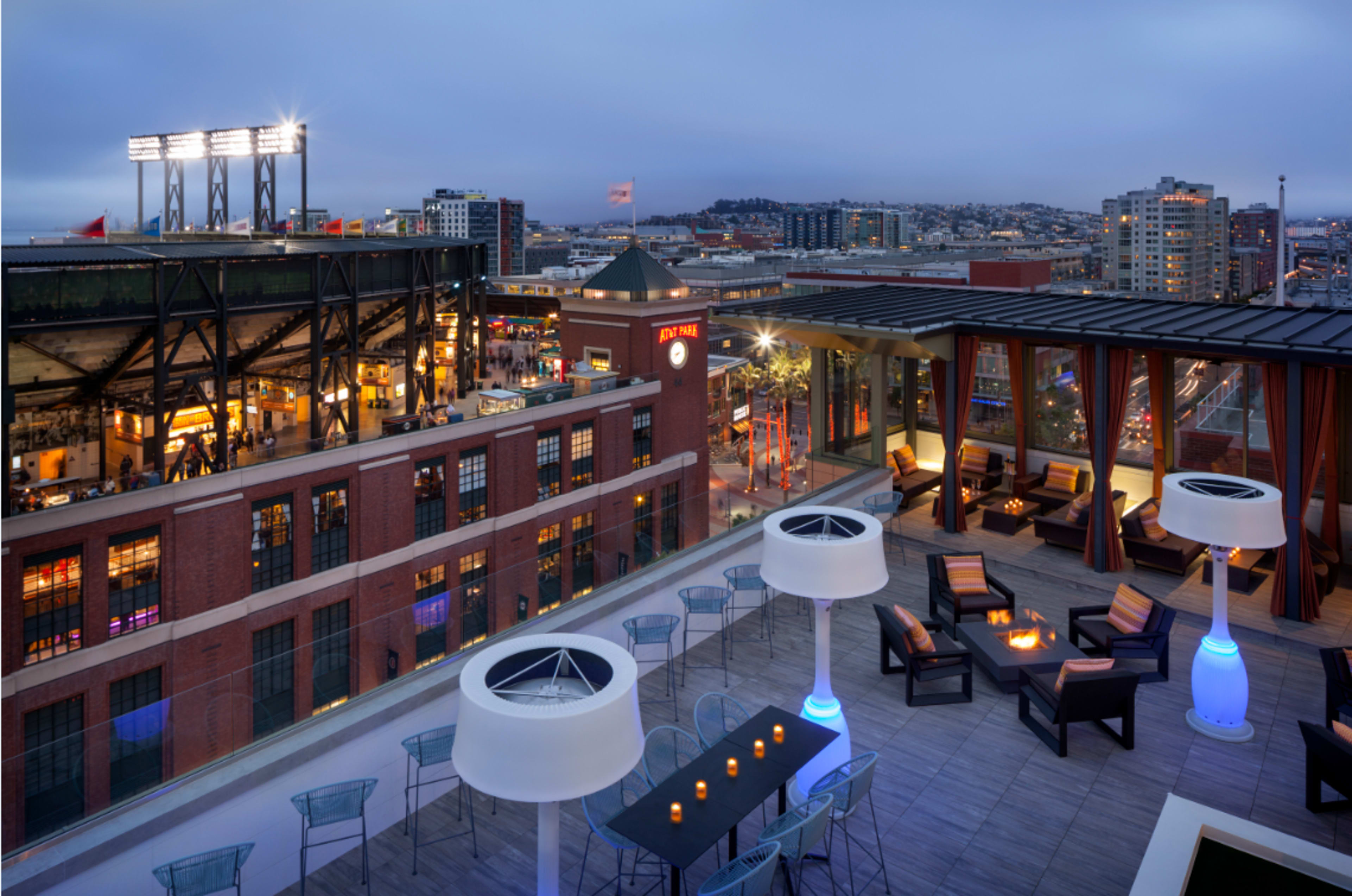 A rooftop lounge overlooking a cityscape that includes a stadium and nearby buildings, with modern seating arrangements and ambient lighting.