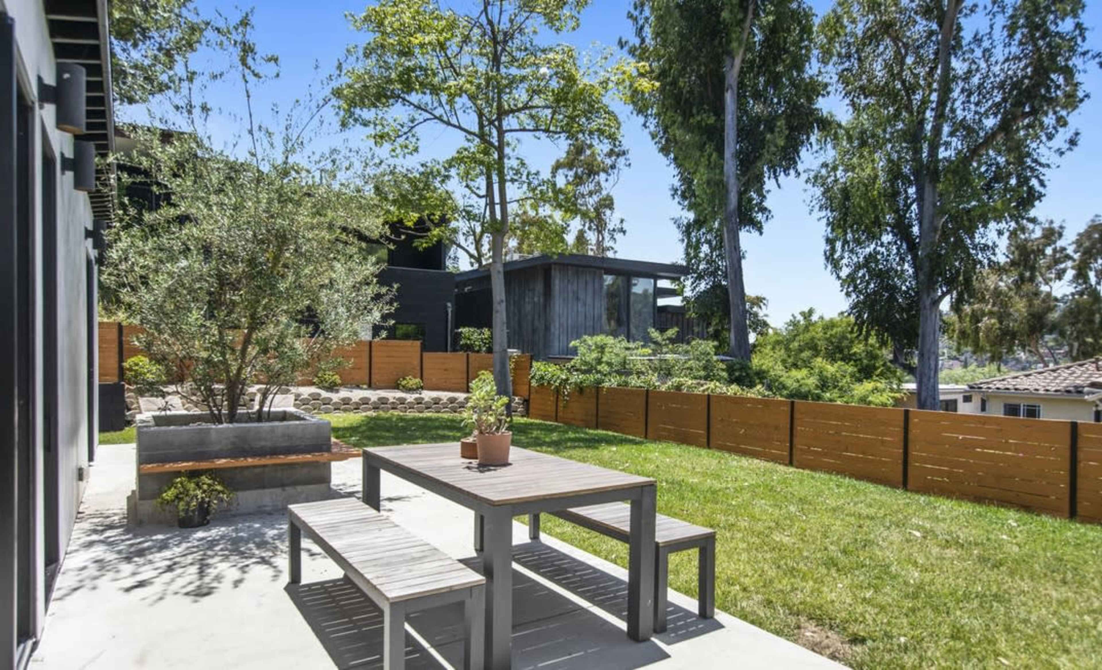 A wooden table and chairs sit on a patio surrounded by grass and trees, with a modern house and a wooden fence in the background.
