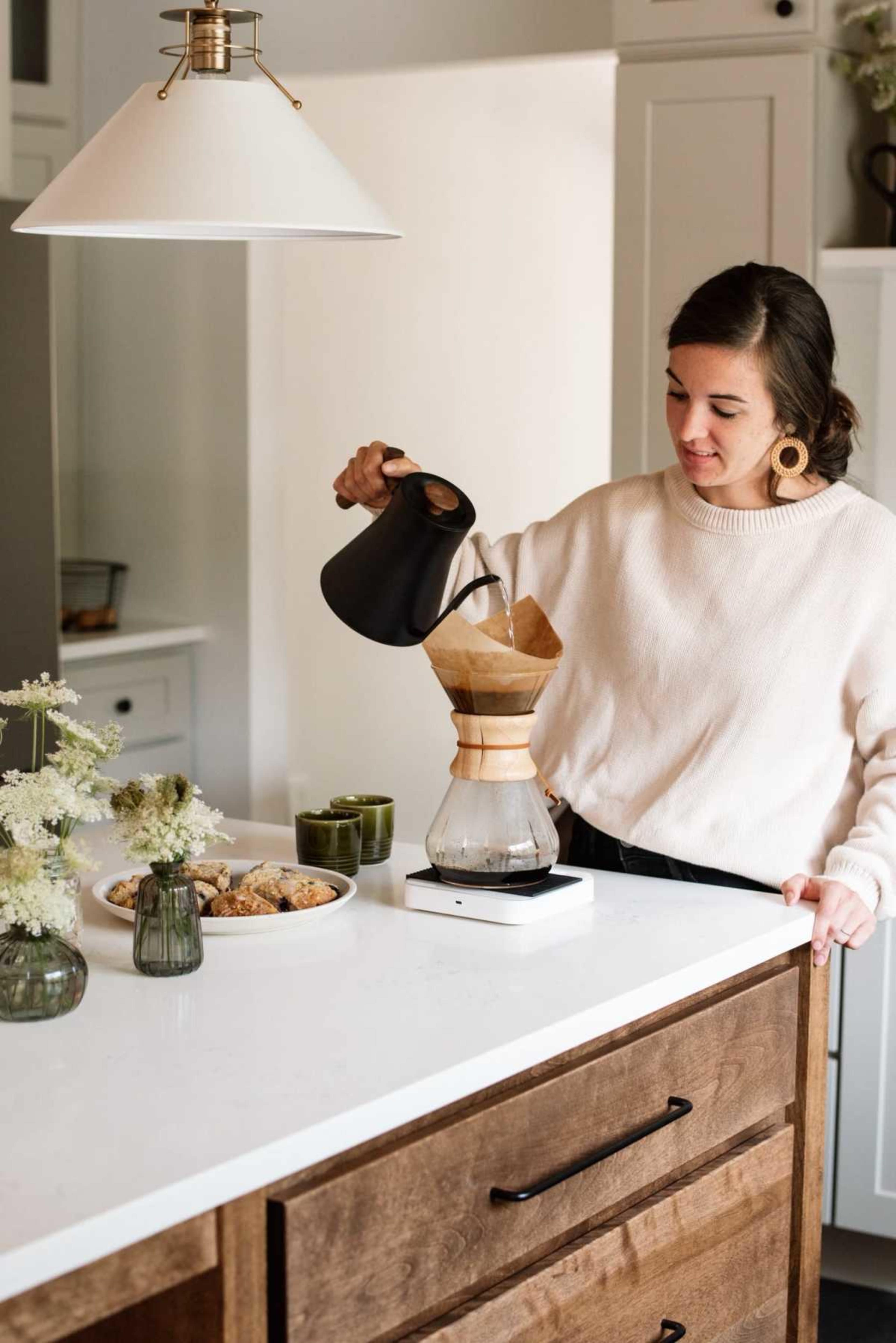 A woman pours hot water into a coffee filter placed in a brew station on a kitchen countertop while surrounded by snacks and small vases of flowers.