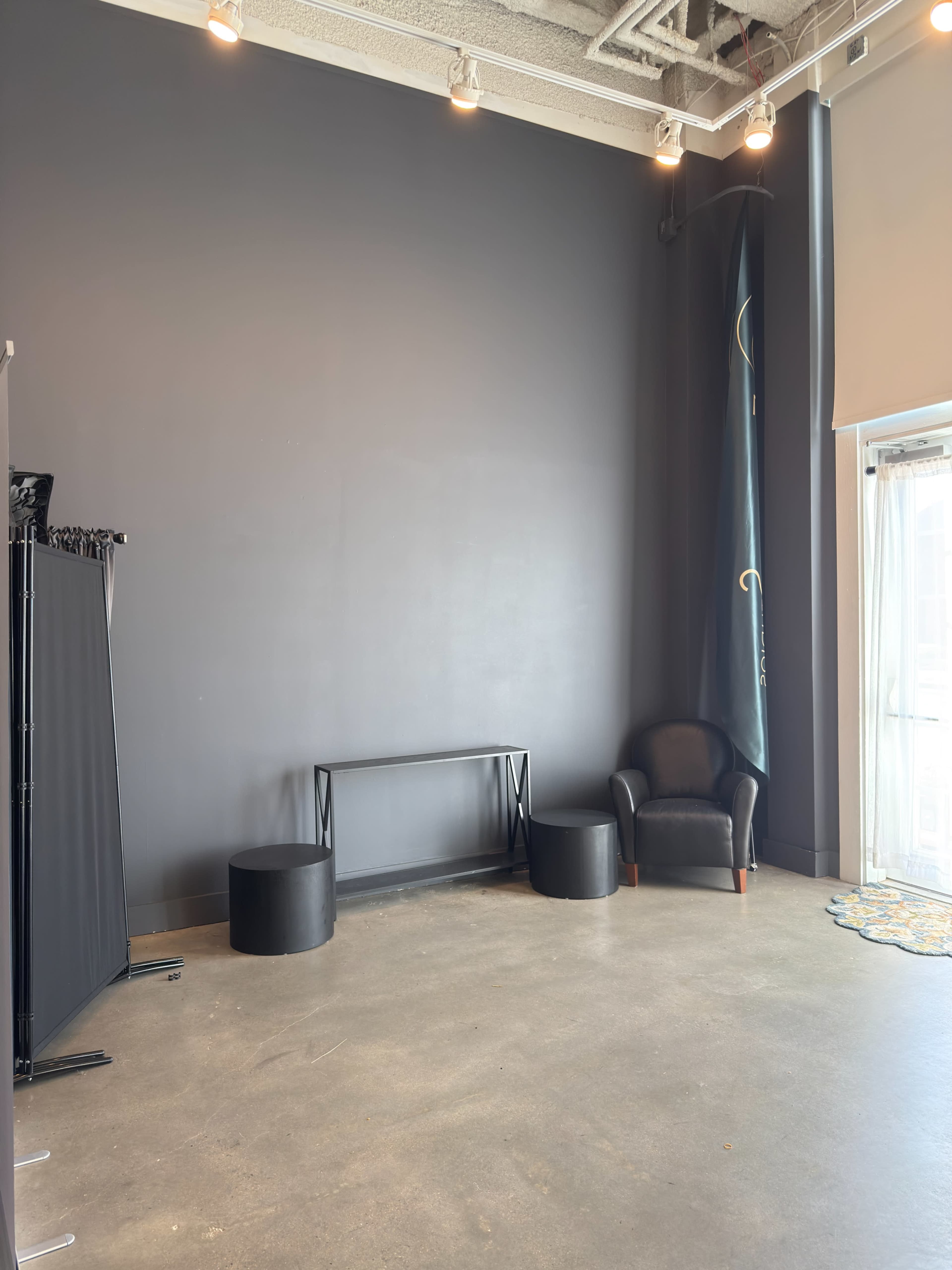 The image shows a spacious, empty room with a dark wall, a black chair, two round black tables, and a window with sheer curtains.