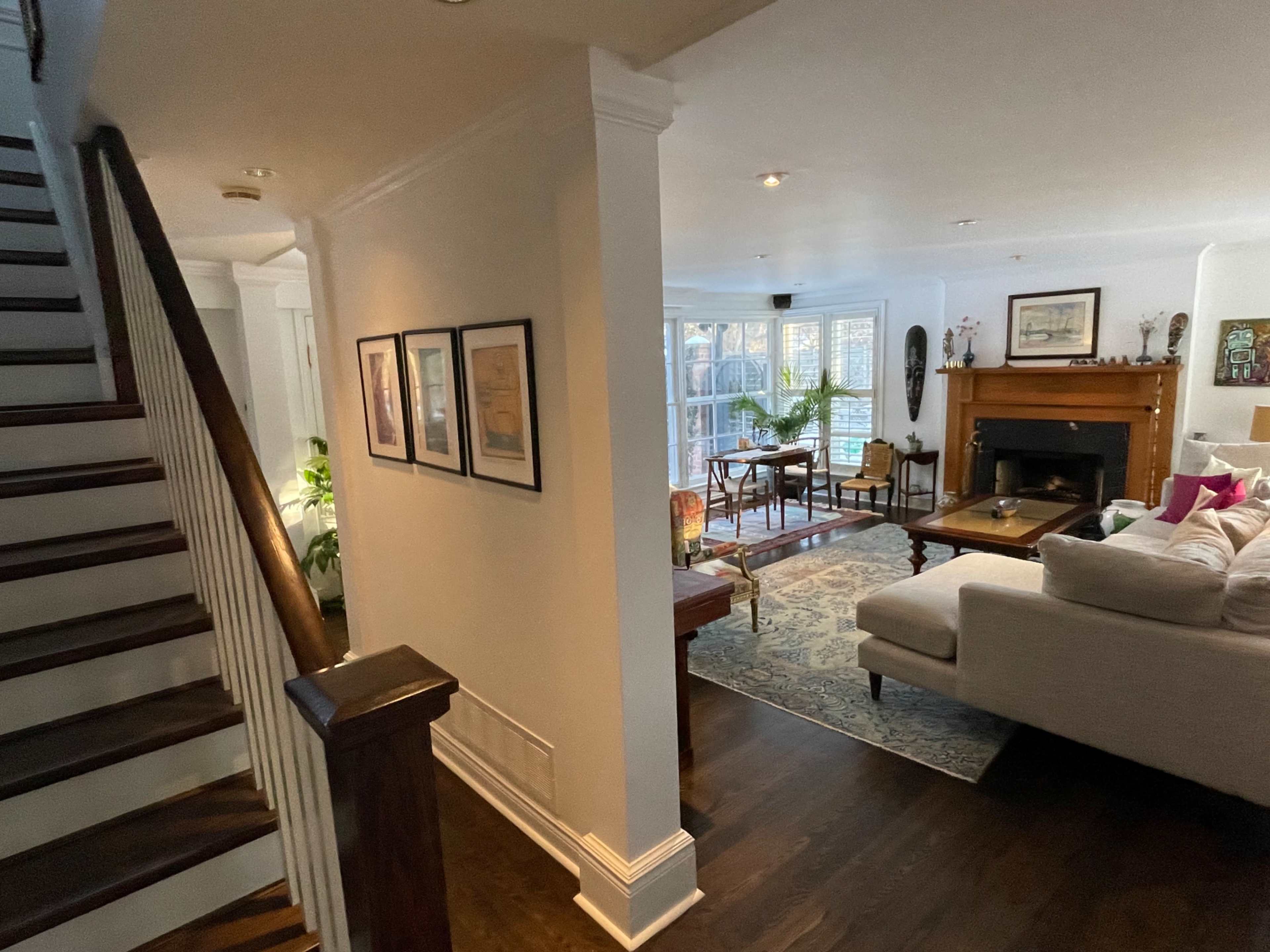 A well-lit living room with a staircase on the left, framed artwork on the wall, and furniture arranged around a fireplace.