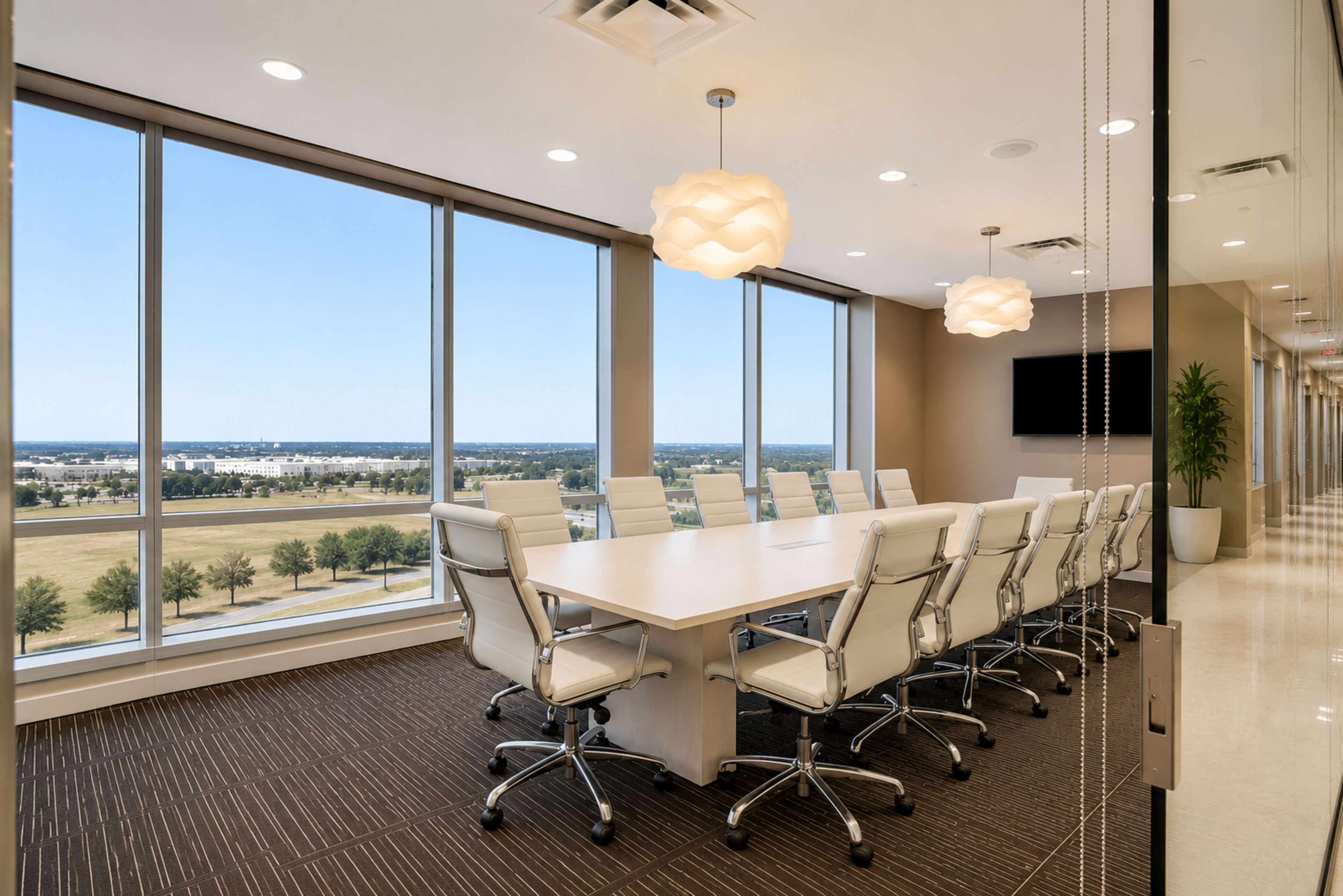 The image shows a modern conference room with a large table and ergonomic chairs, surrounded by floor-to-ceiling windows that offer a view of the landscape outside.