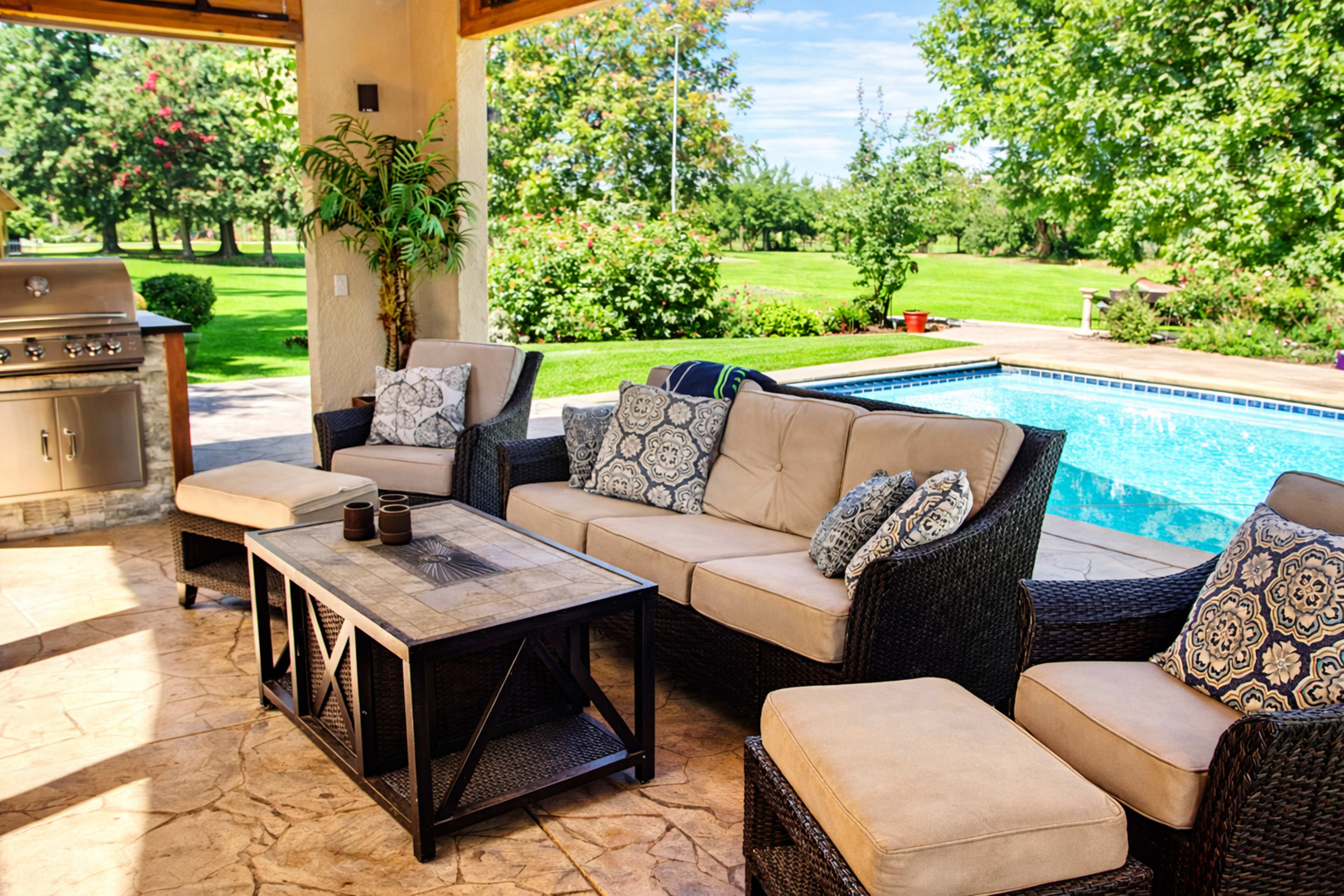 A patio area features a seating arrangement with beige cushions, a coffee table, and a grill nearby, overlooking a swimming pool and lush greenery.