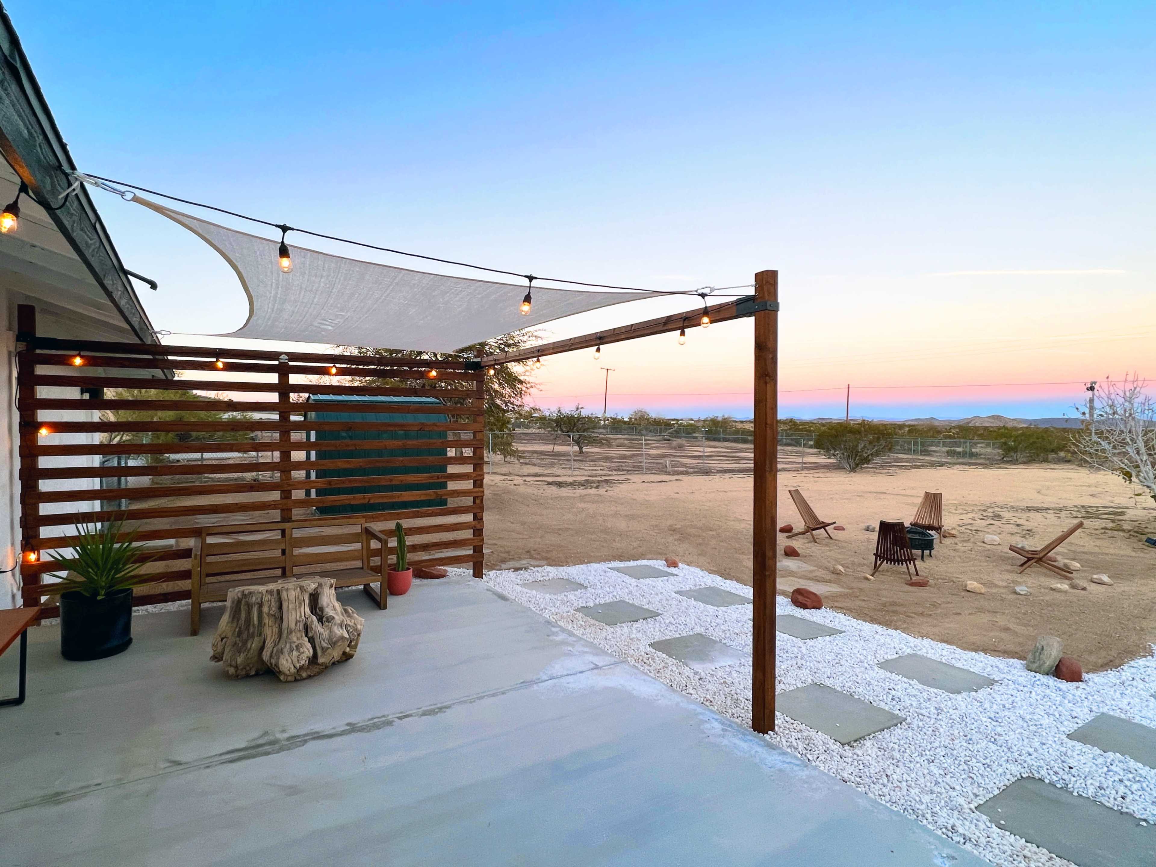 A wooden patio area features a shade sail and a fire pit surrounded by gravel, with desert terrain visible in the background at sunset.