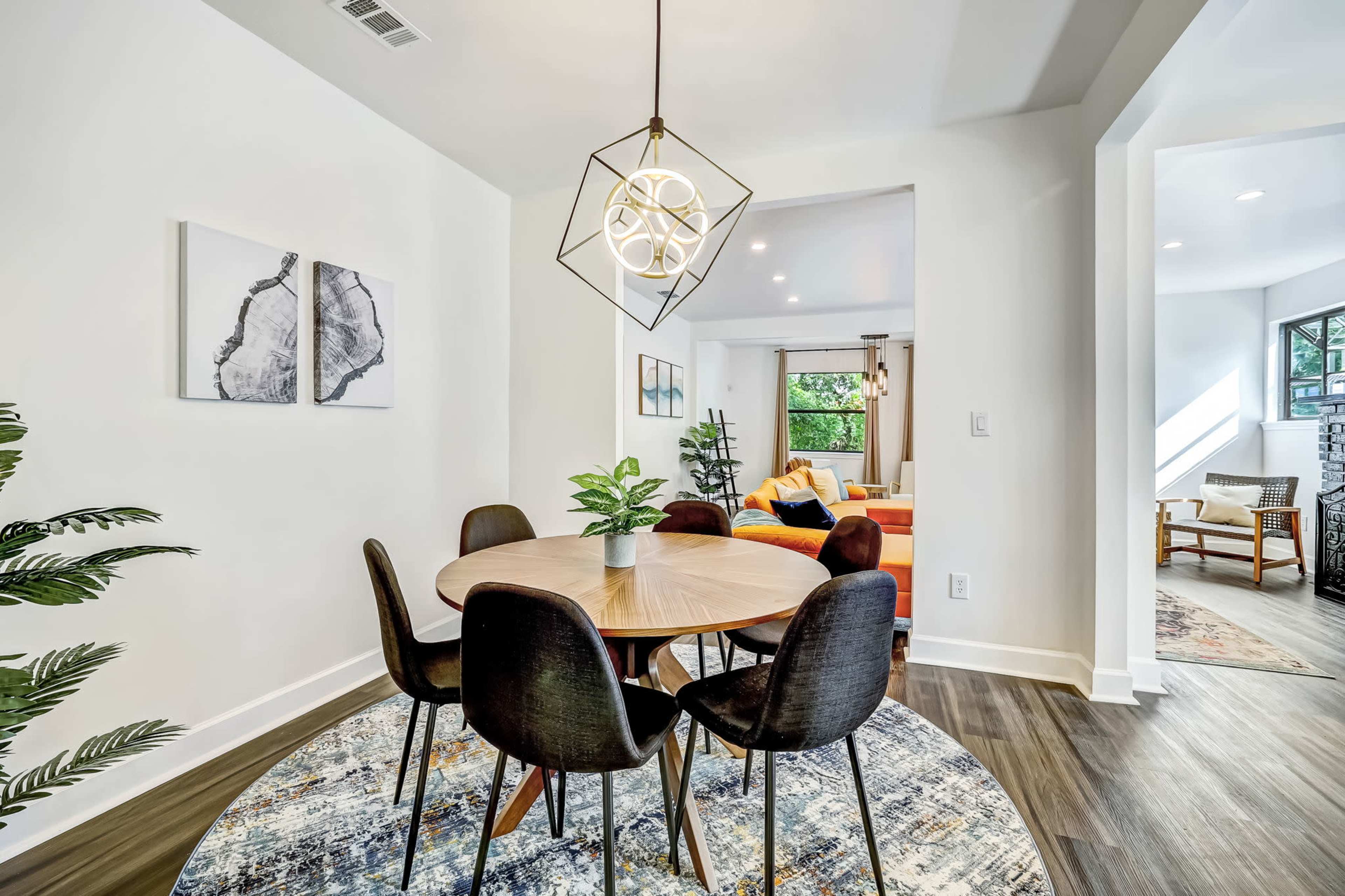 A modern dining area features a round wooden table surrounded by black chairs, with a decorative plant and artistic wall prints.