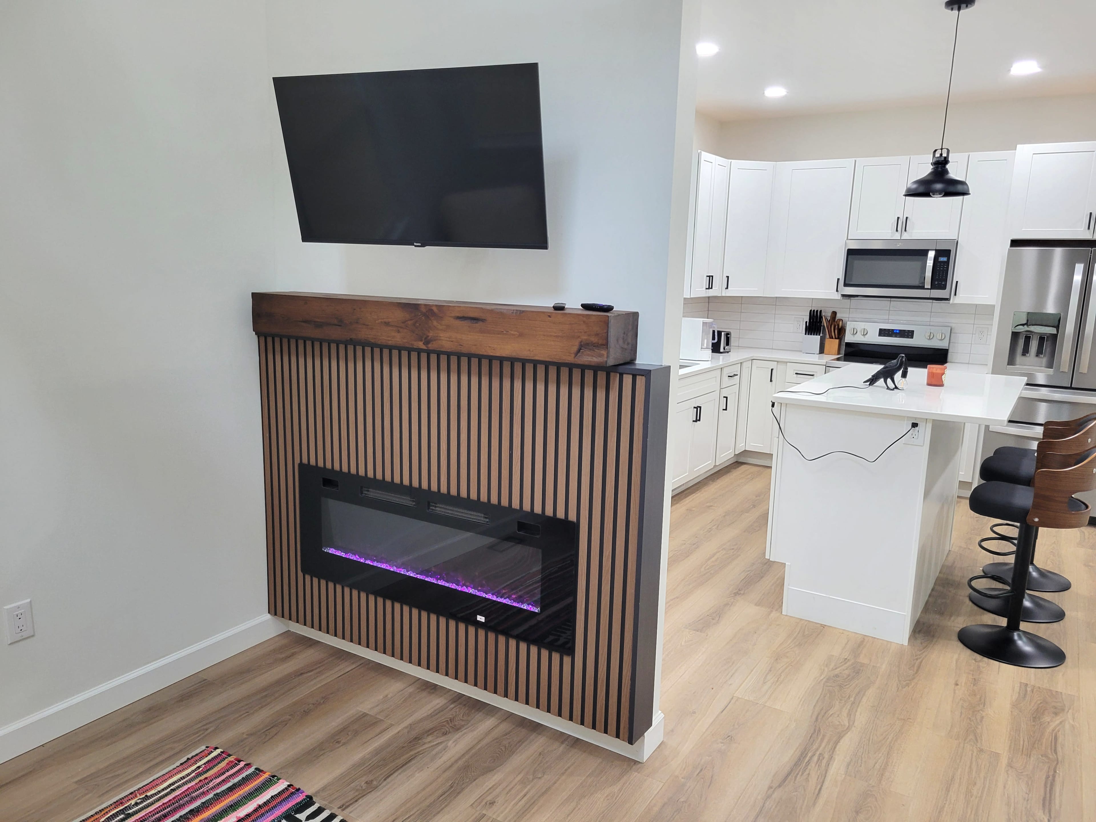The image shows a modern living space featuring a wall-mounted television above a linear electric fireplace with a wooden mantle and vertical slats, adjacent to a kitchen area with white cabinetry and black bar stools at a counter.