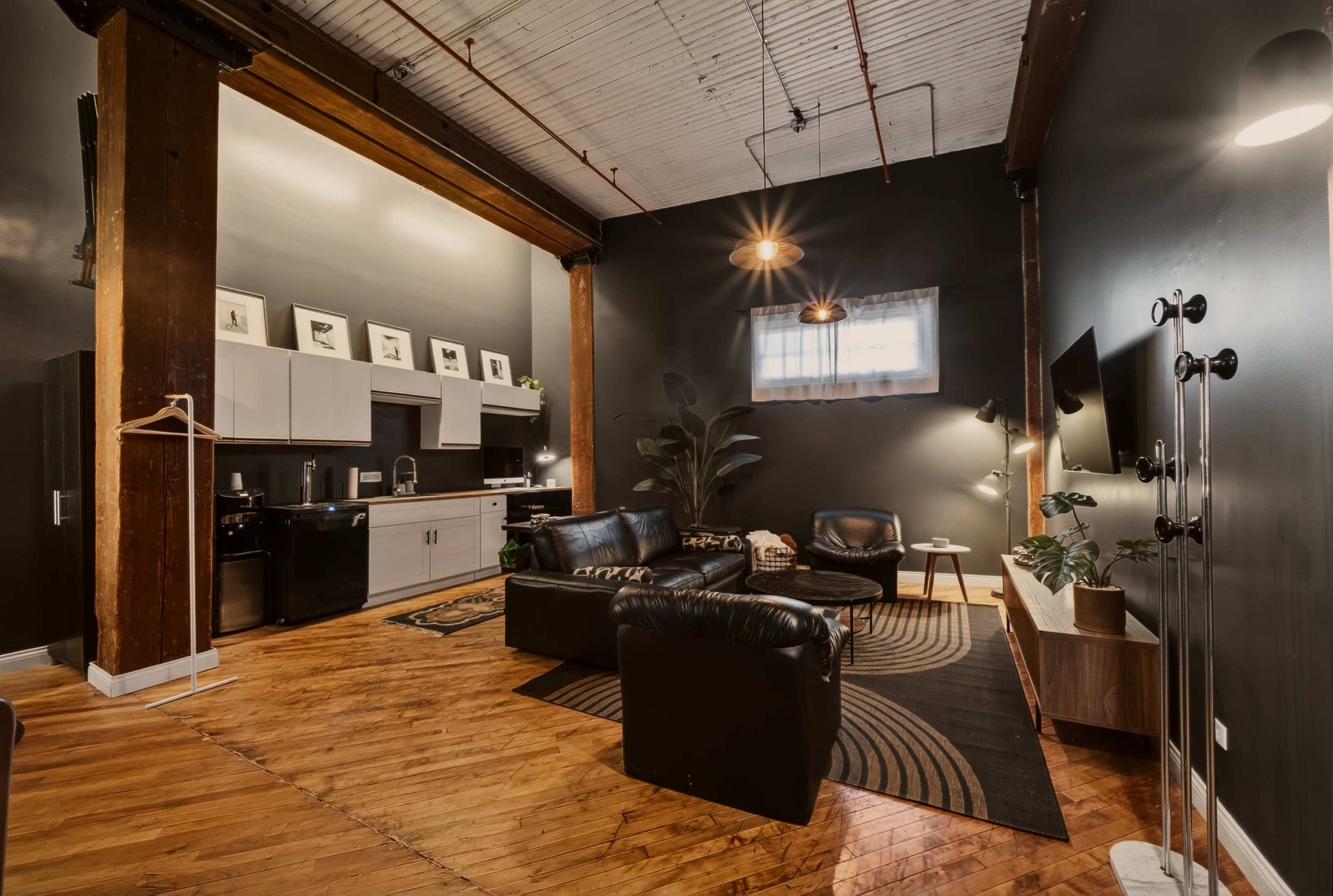 The image shows a modern living space featuring dark walls, wooden beams, a kitchenette, a seating area with black leather furniture, and decorative plants.