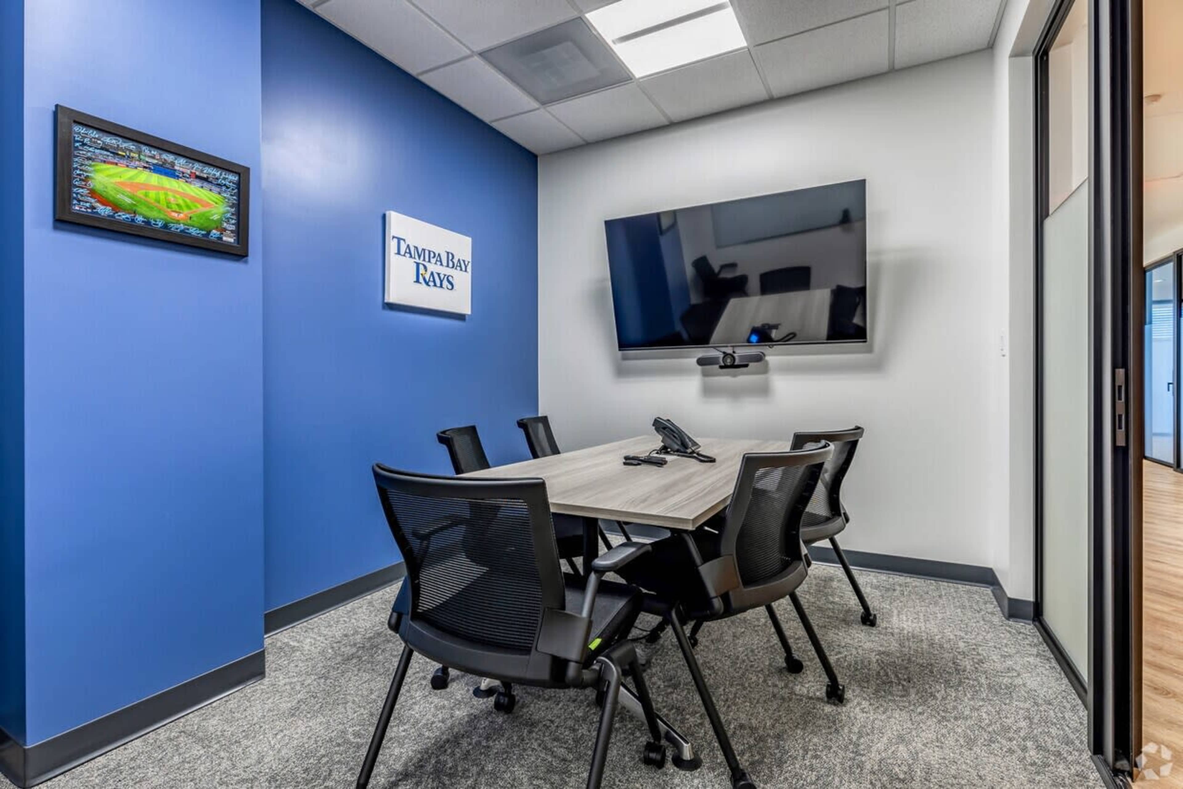 A small conference room features a round table, six black chairs, a large TV mounted on the wall, and a logo of the Tampa Bay Rays.