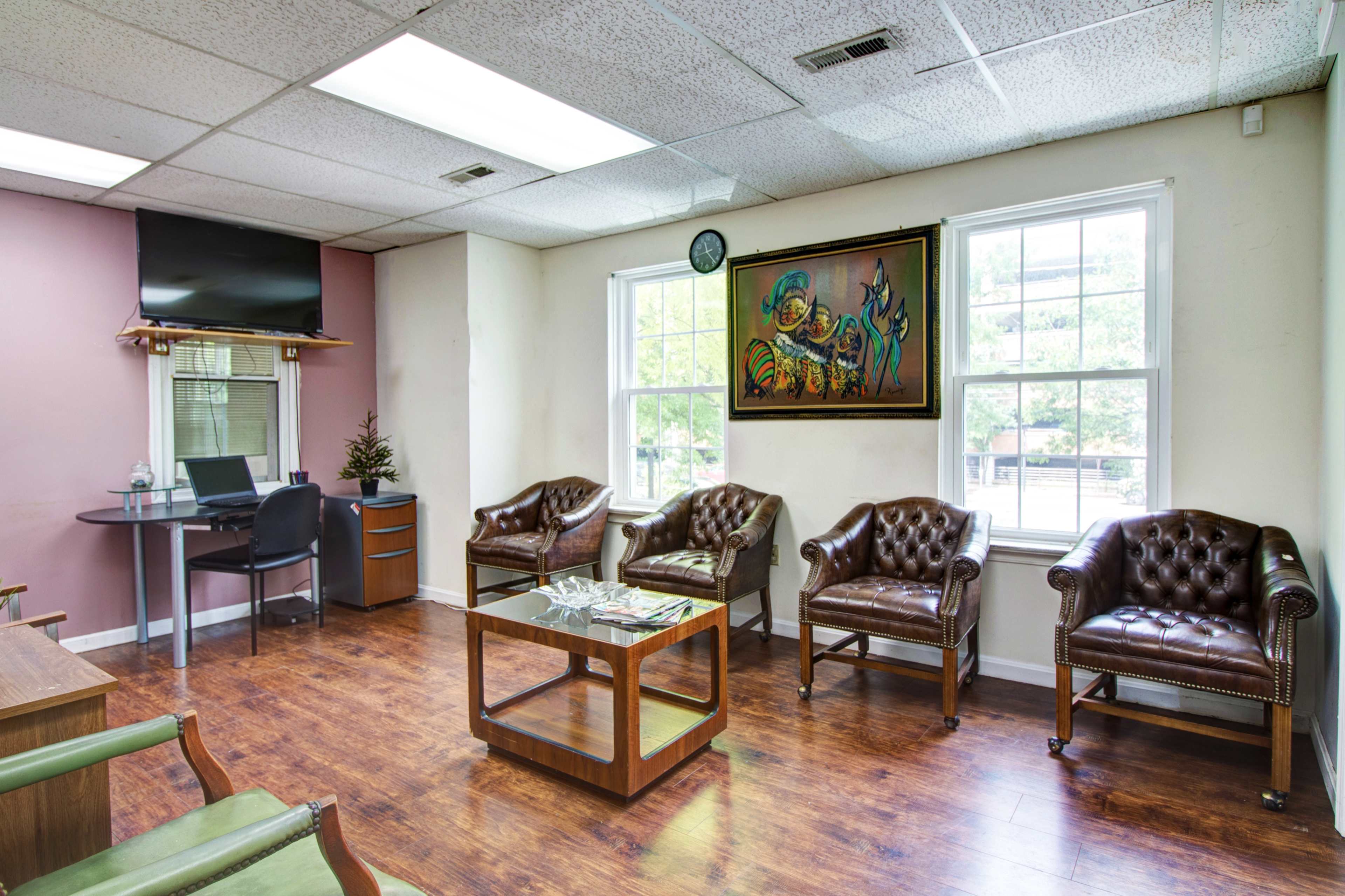 A waiting room with brown leather chairs, a small desk with a monitor, and a colorful painting on the wall.