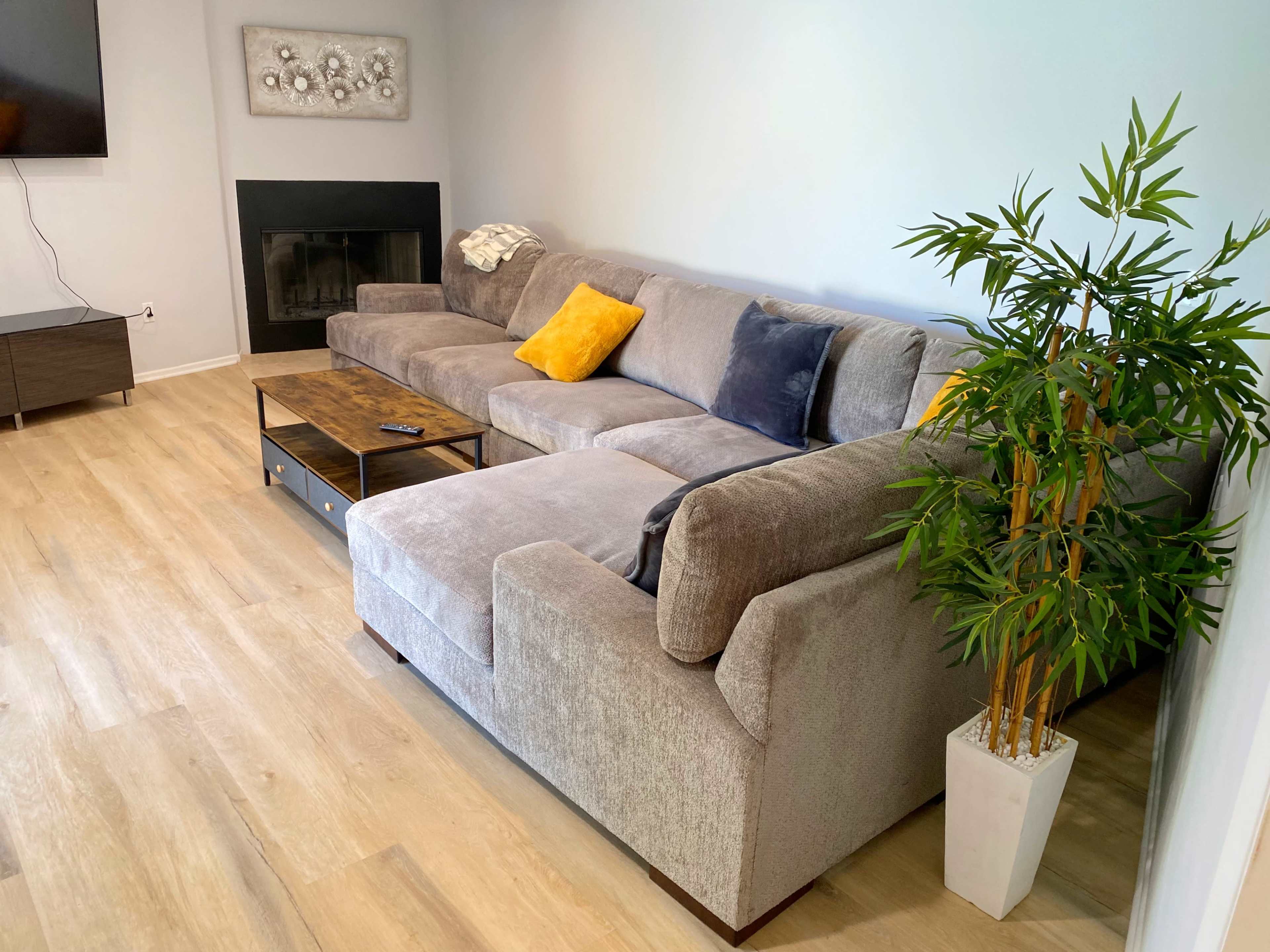 A modern living room with a grey sectional sofa, a wooden coffee table, a small potted plant, and a wall-mounted television.