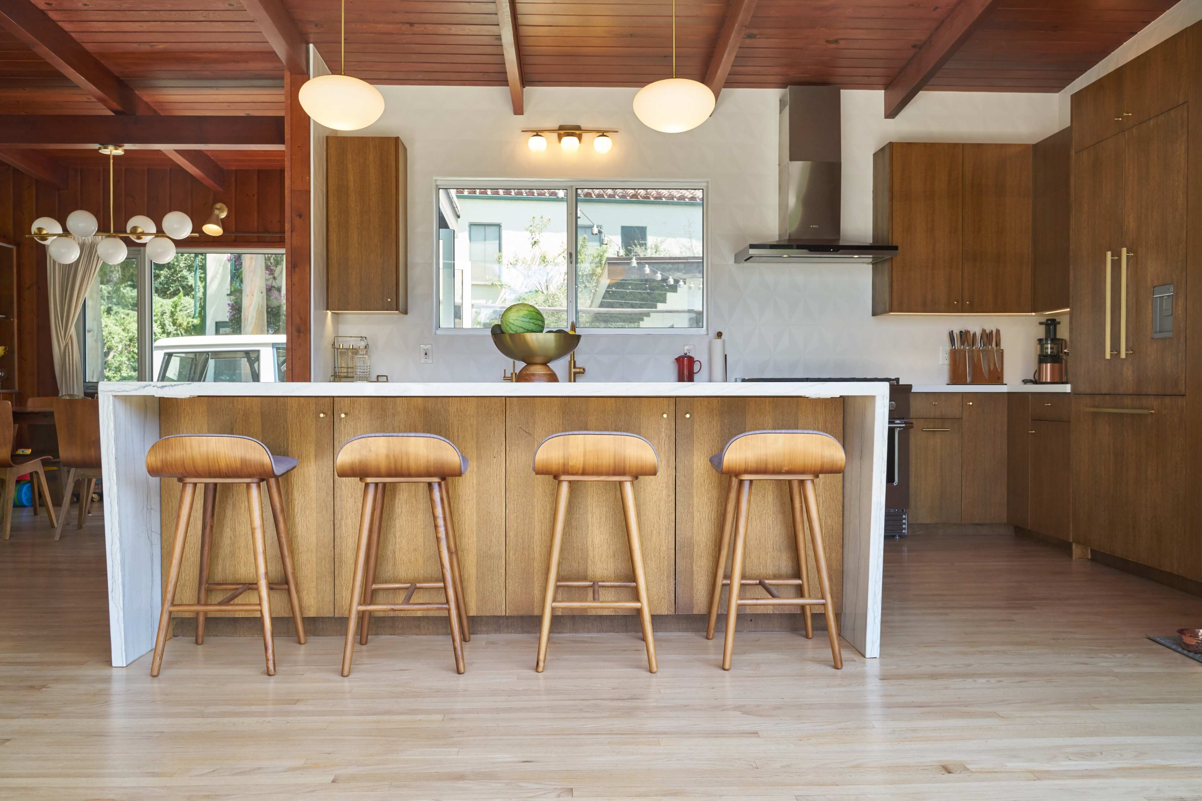 A modern kitchen featuring a central island with four wooden bar stools, wooden cabinetry, and a large window allowing natural light.
