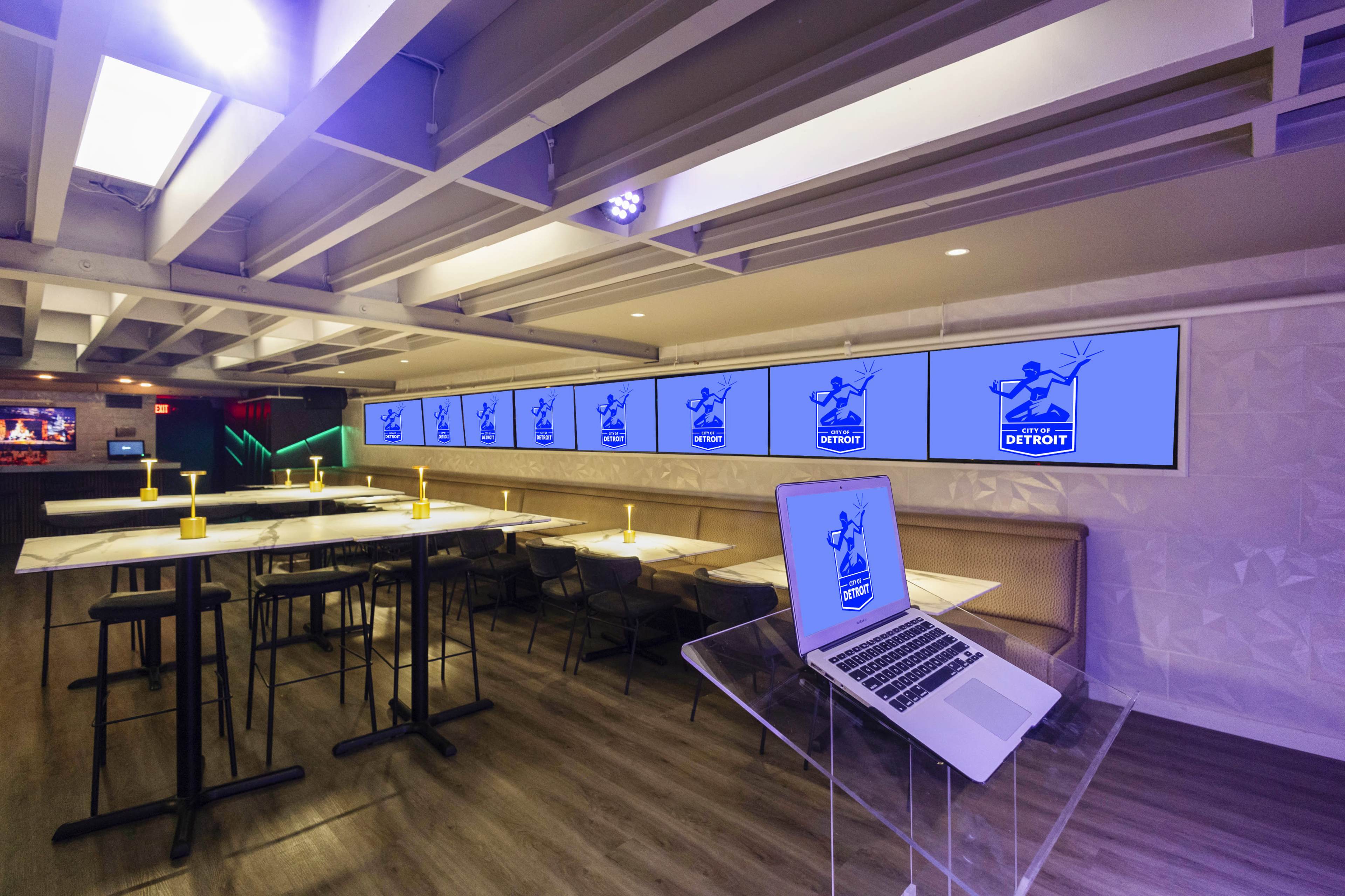The image shows a modern dining area with high tables, chairs, and multiple screens displaying graphics along the wall.