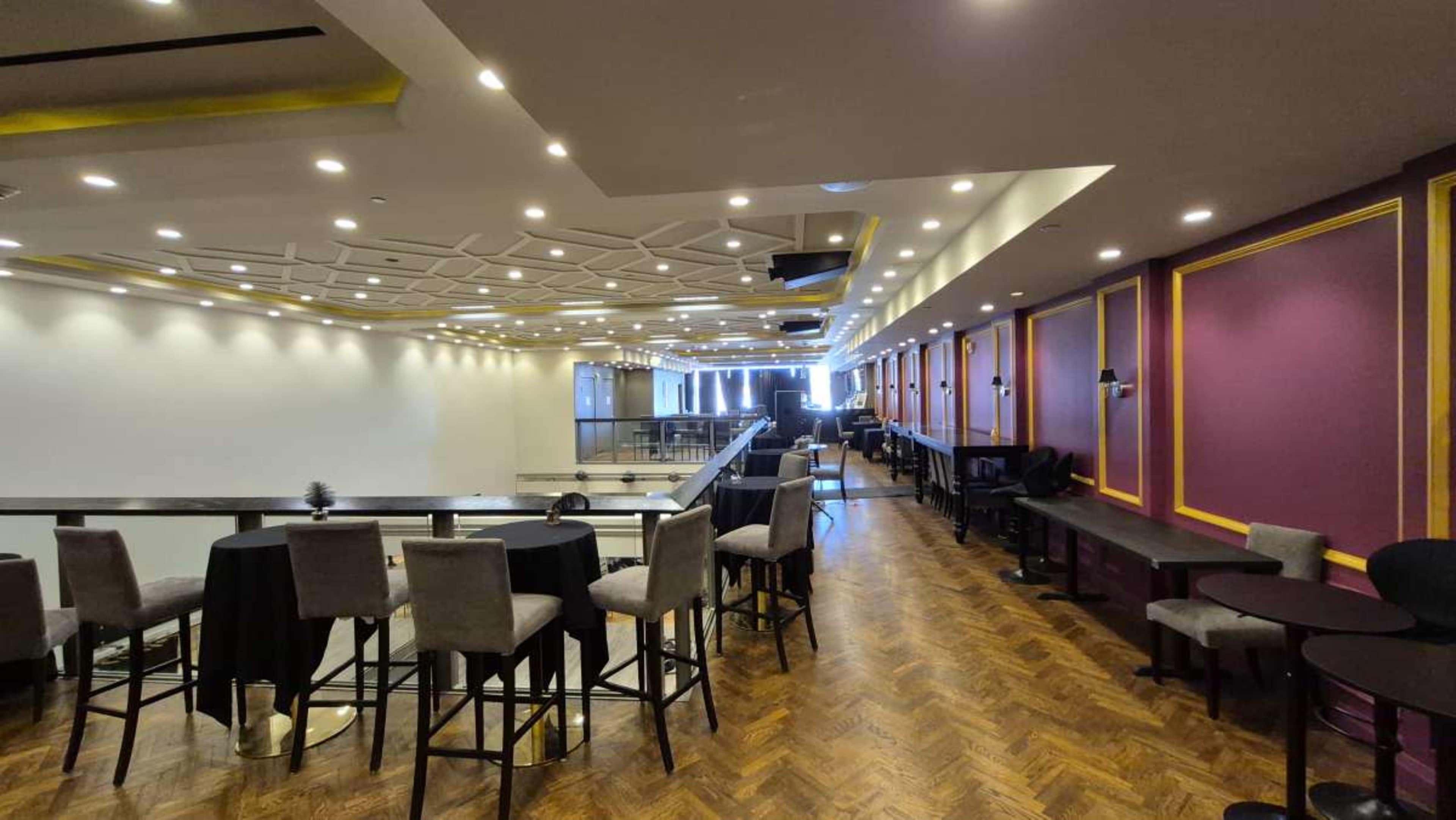 The image shows a modern event space with a mix of tall tables and chairs, stylish lighting, and a polished wooden floor.