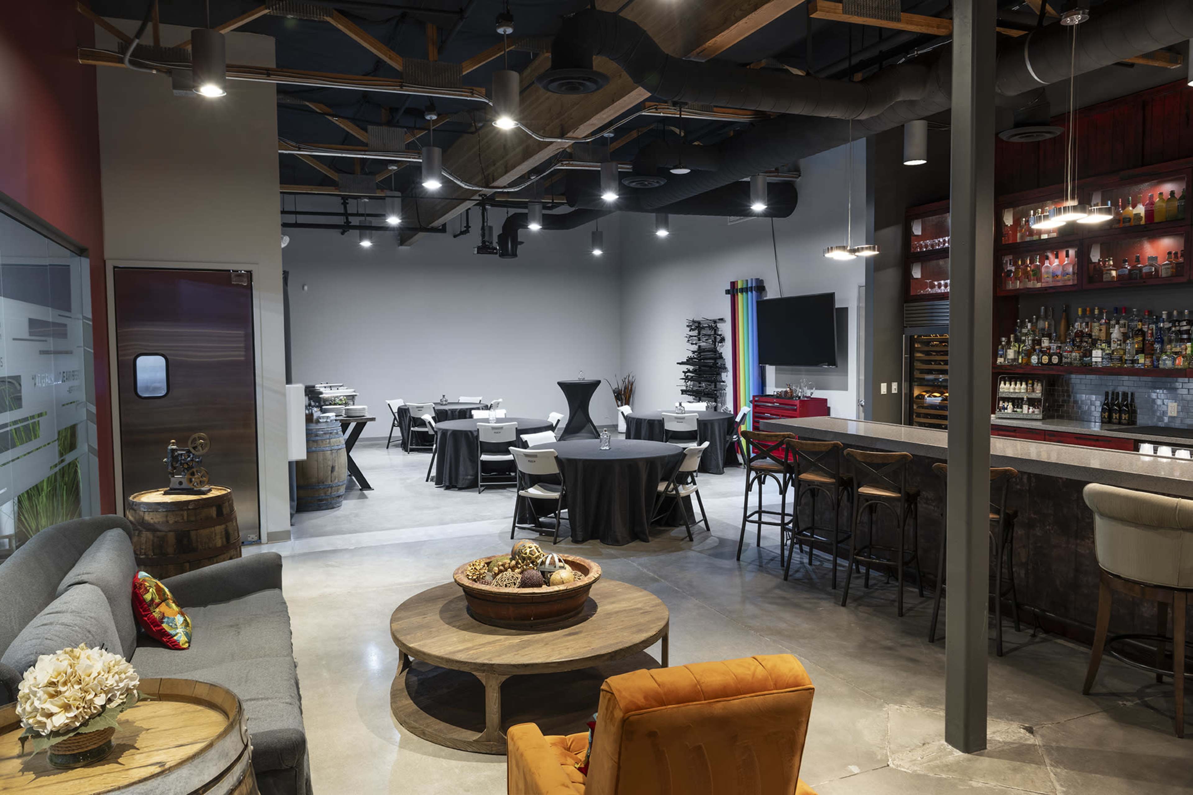 The image shows a modern interior of a restaurant or event space with a seating area featuring a sofa and decorative table, along with dining tables set up for an event in the background.