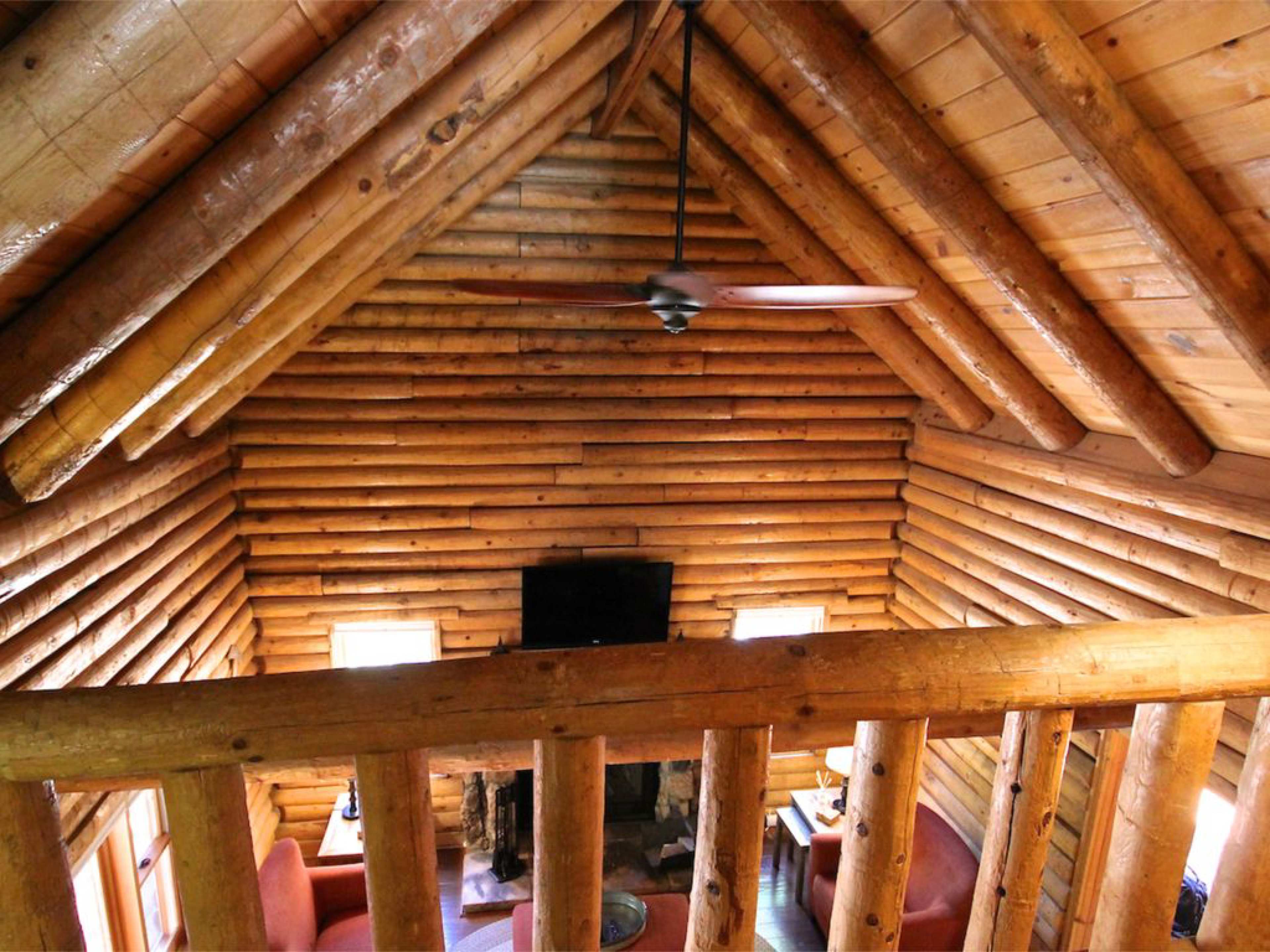 The interior of a log cabin features a high ceiling with wooden beams, a ceiling fan, and a seating area with two chairs in front of a stone fireplace.