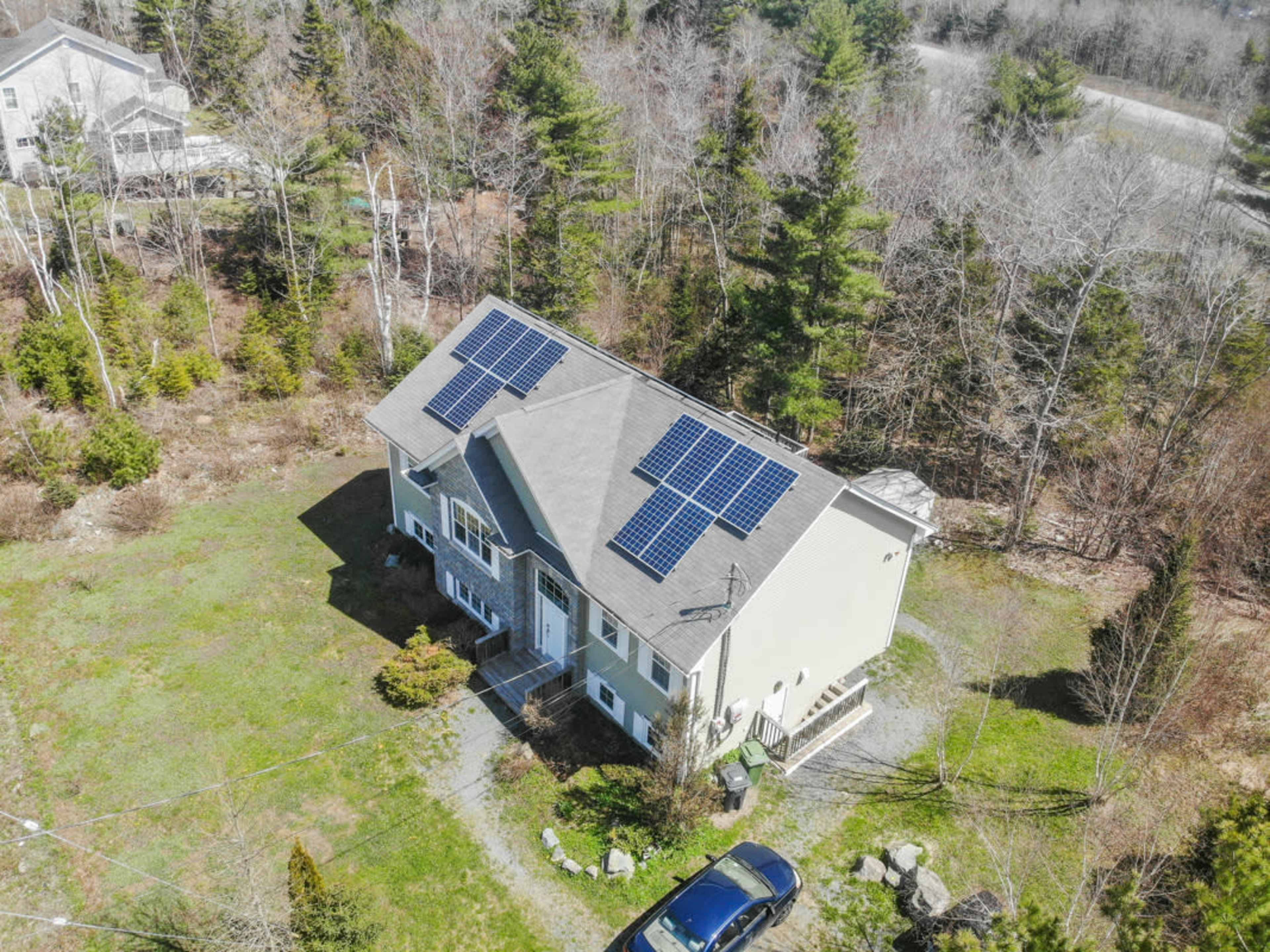 A two-story house with solar panels on the roof is situated on a grassy lot surrounded by trees.