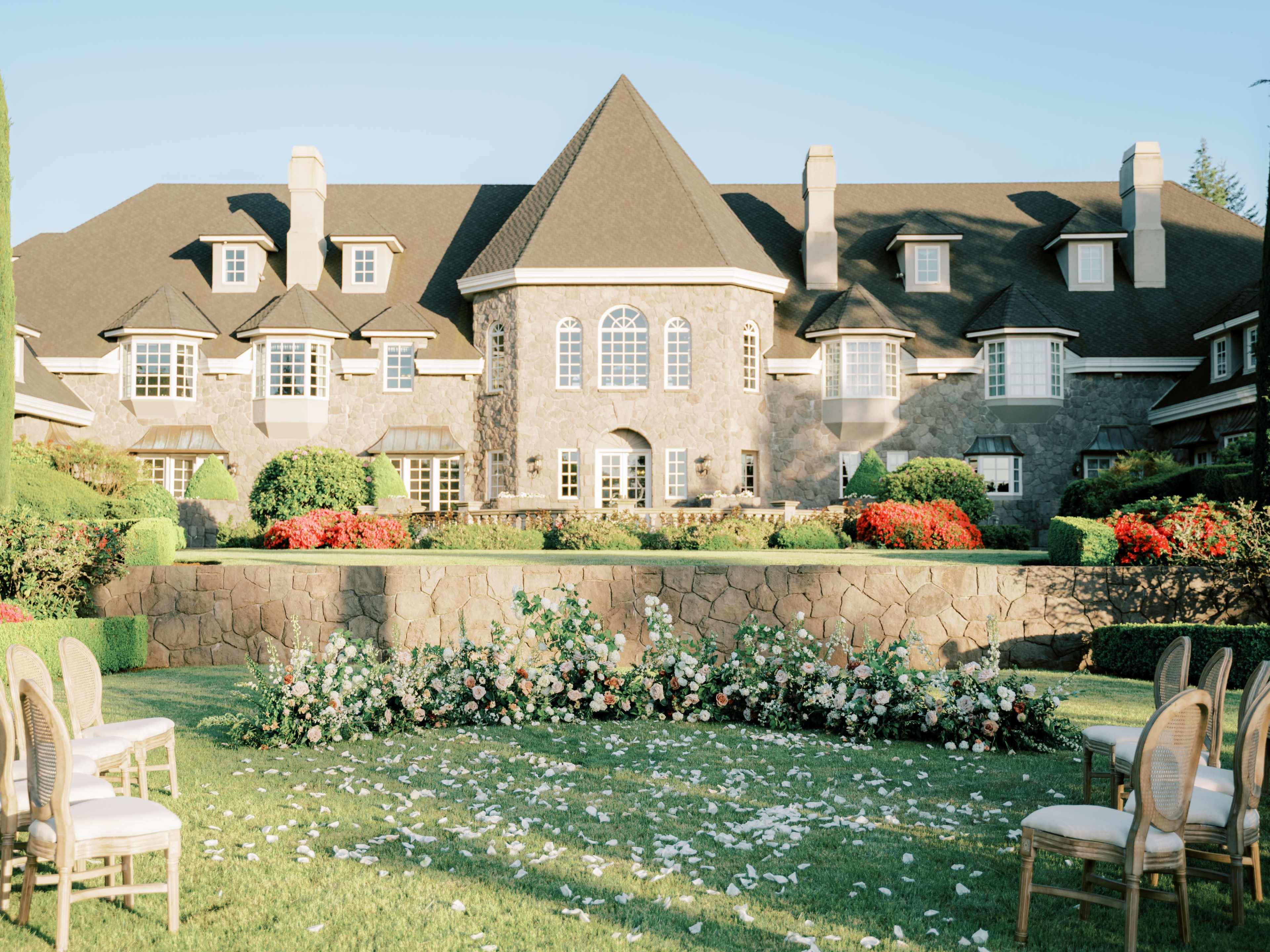 A large stone mansion with a landscaped garden featuring floral arrangements and chairs arranged in front of it.