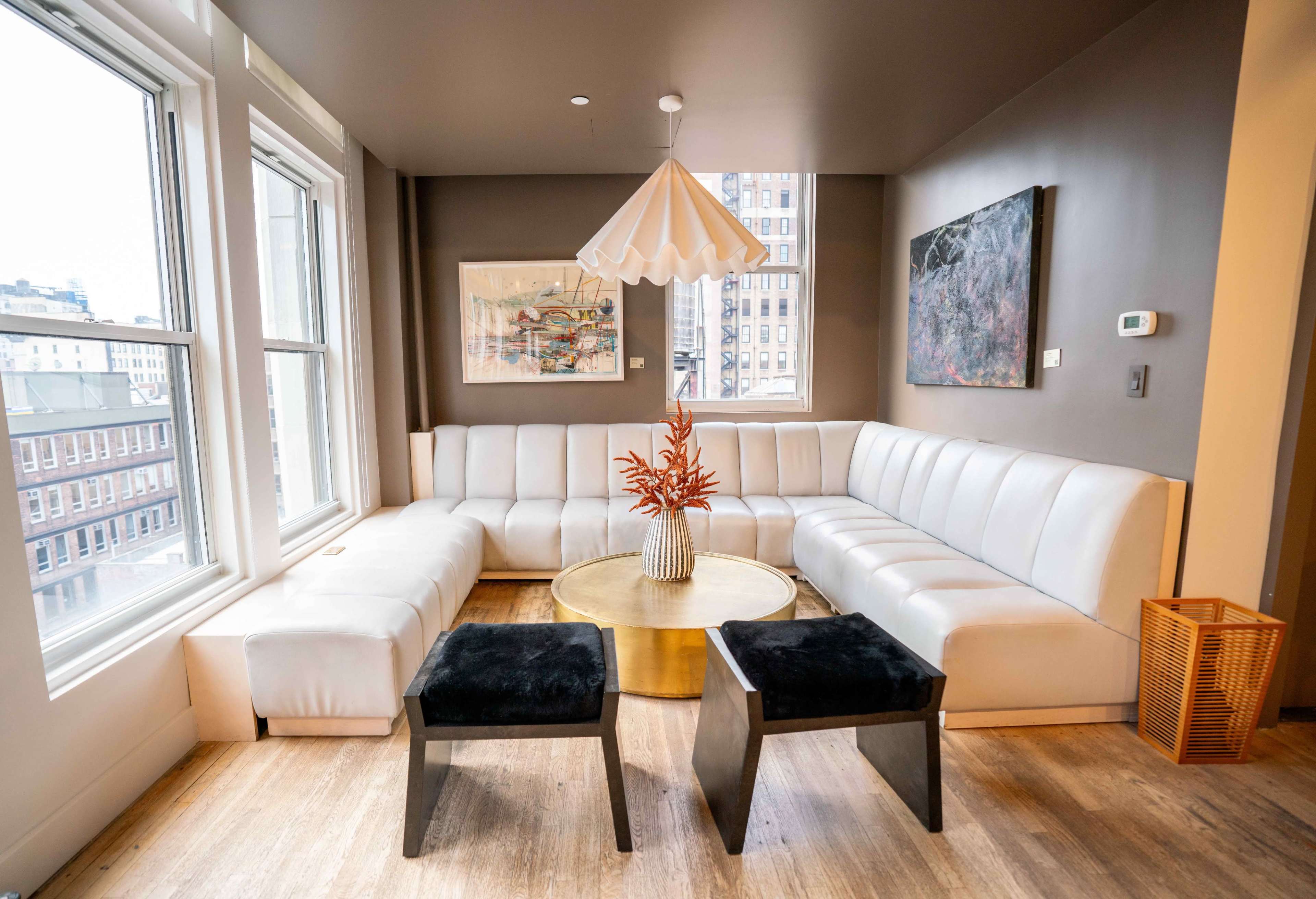The image shows a modern, light-colored sectional sofa arranged in a corner of a room with large windows, a round gold coffee table, and two black stools.