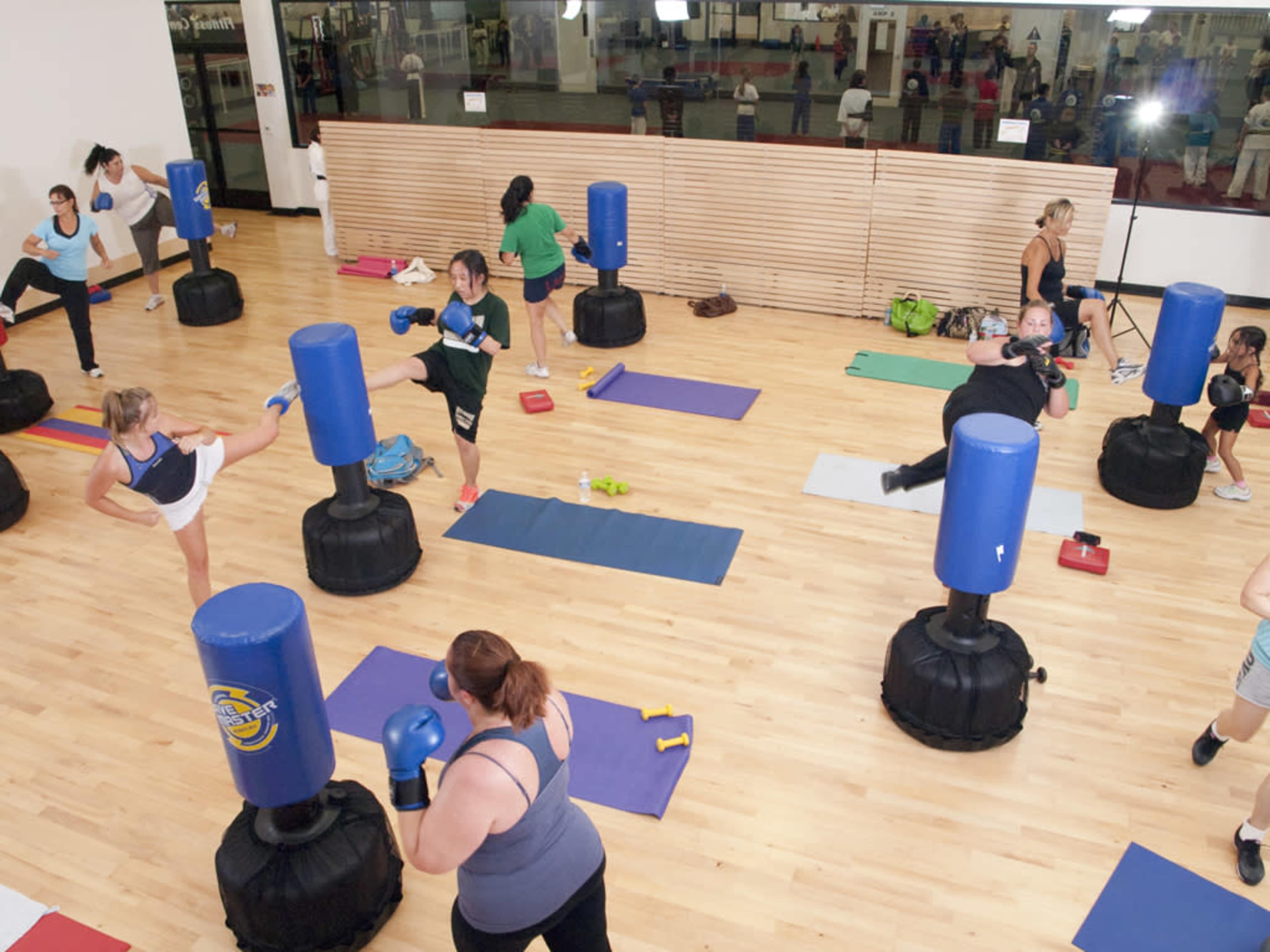 A fitness class is taking place in a gym, with participants engaging in various exercises around blue punching bags on a wooden floor.