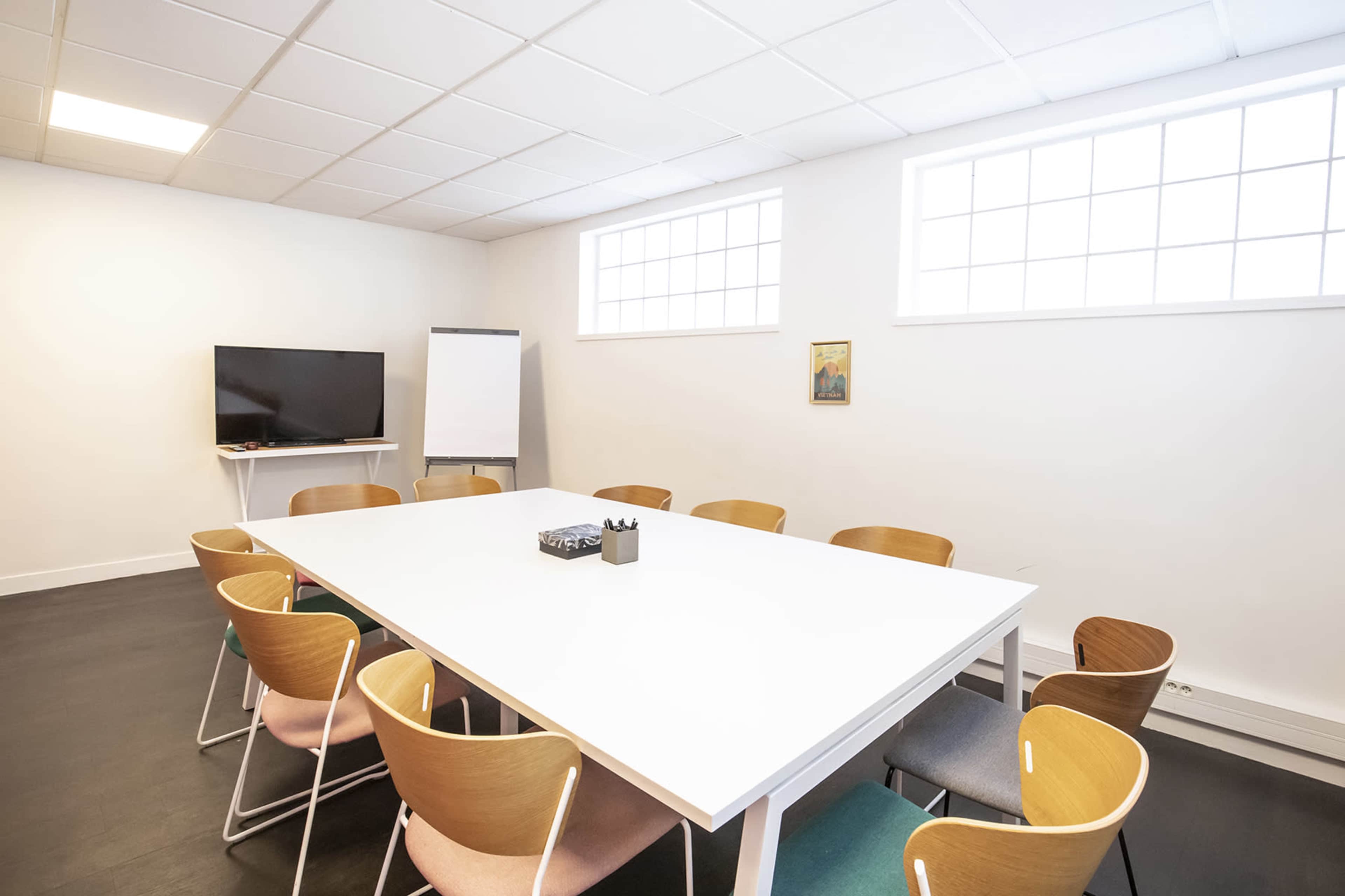 A modern conference room features a large white table surrounded by wooden chairs, with a television and a whiteboard in the corner.
