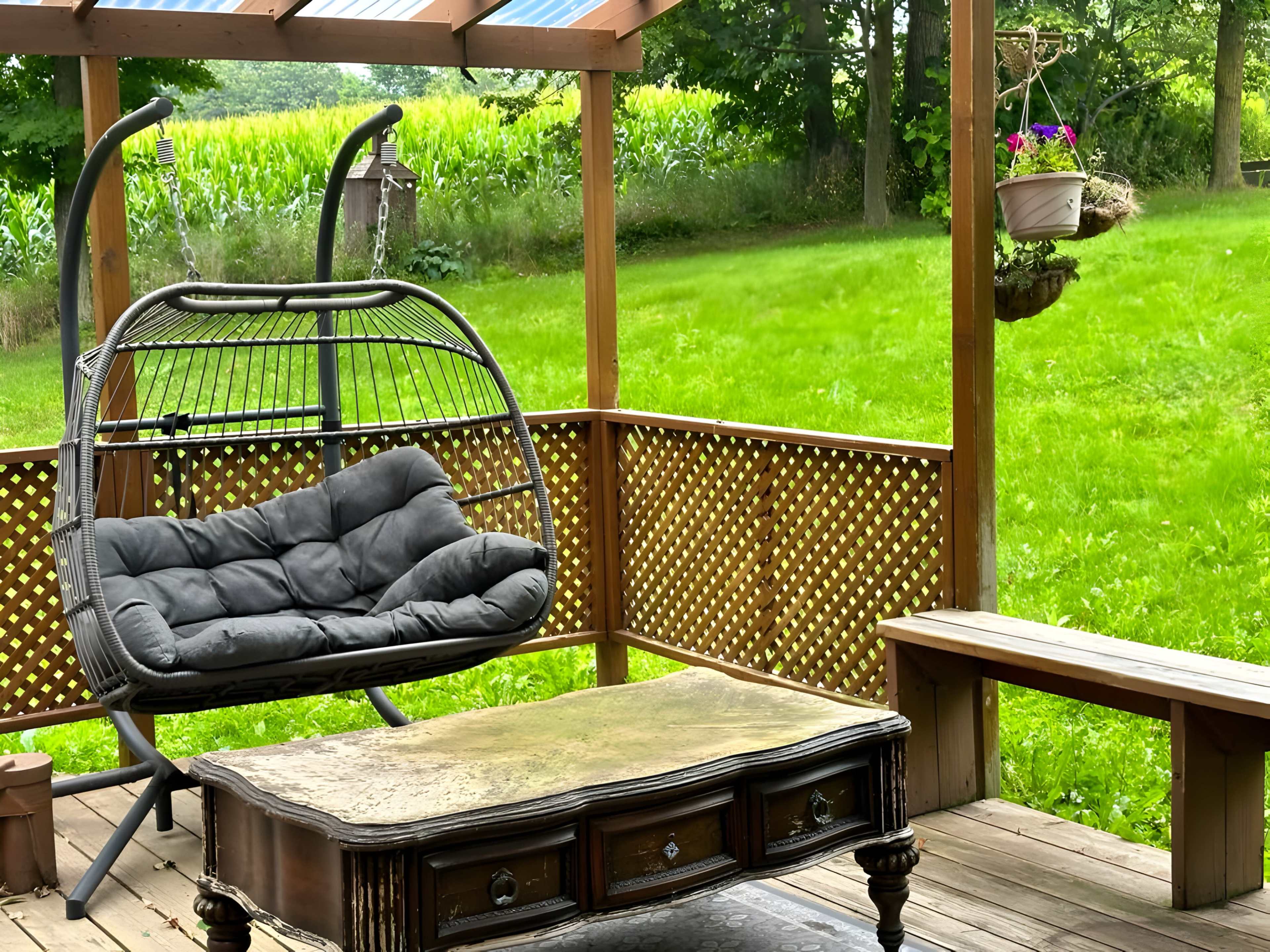 A suspended chair and a wooden coffee table are arranged on a deck surrounded by green grass and trees.