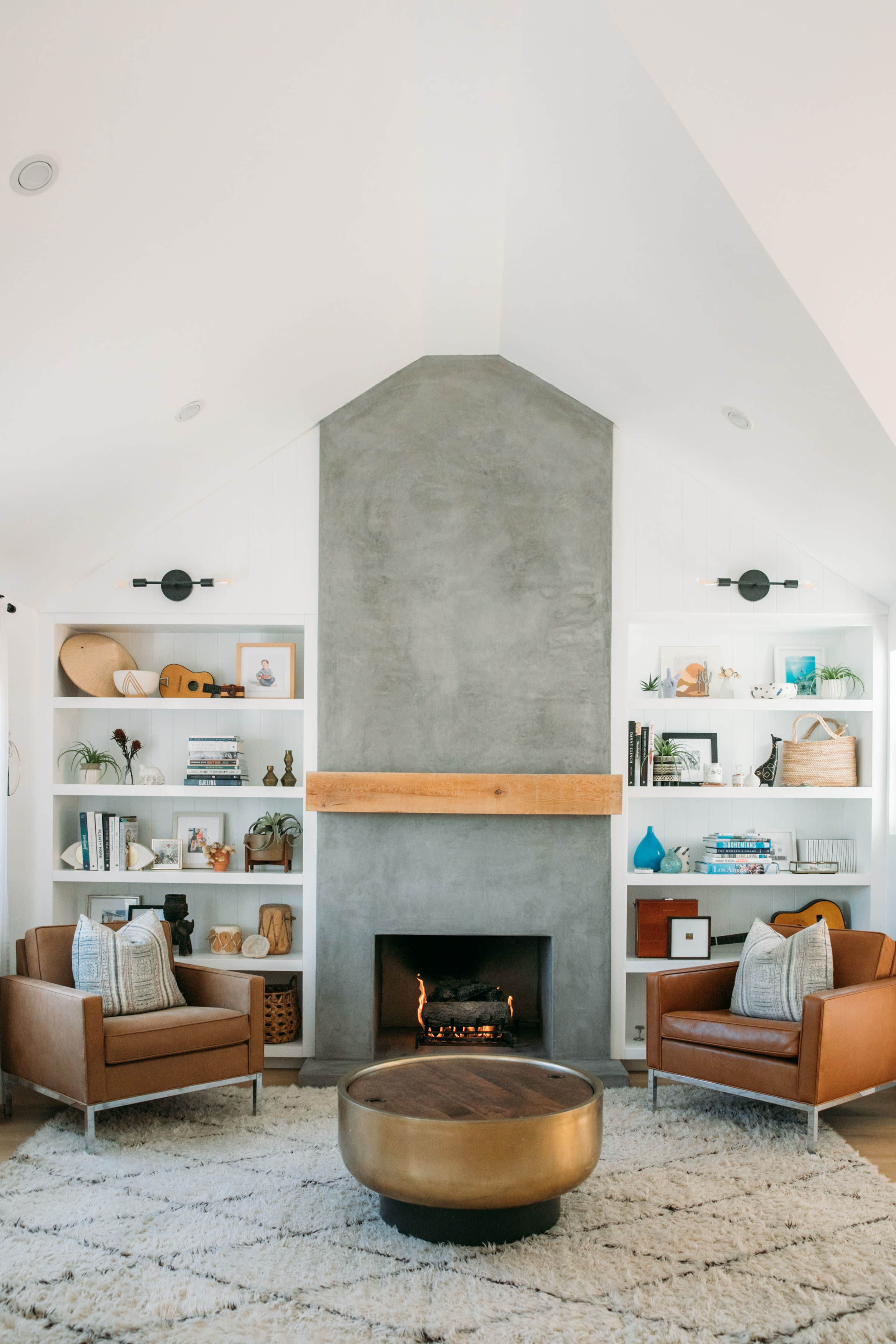 A modern living room features two brown leather chairs facing a round coffee table, with a concrete fireplace and built-in shelves filled with various decor and books.