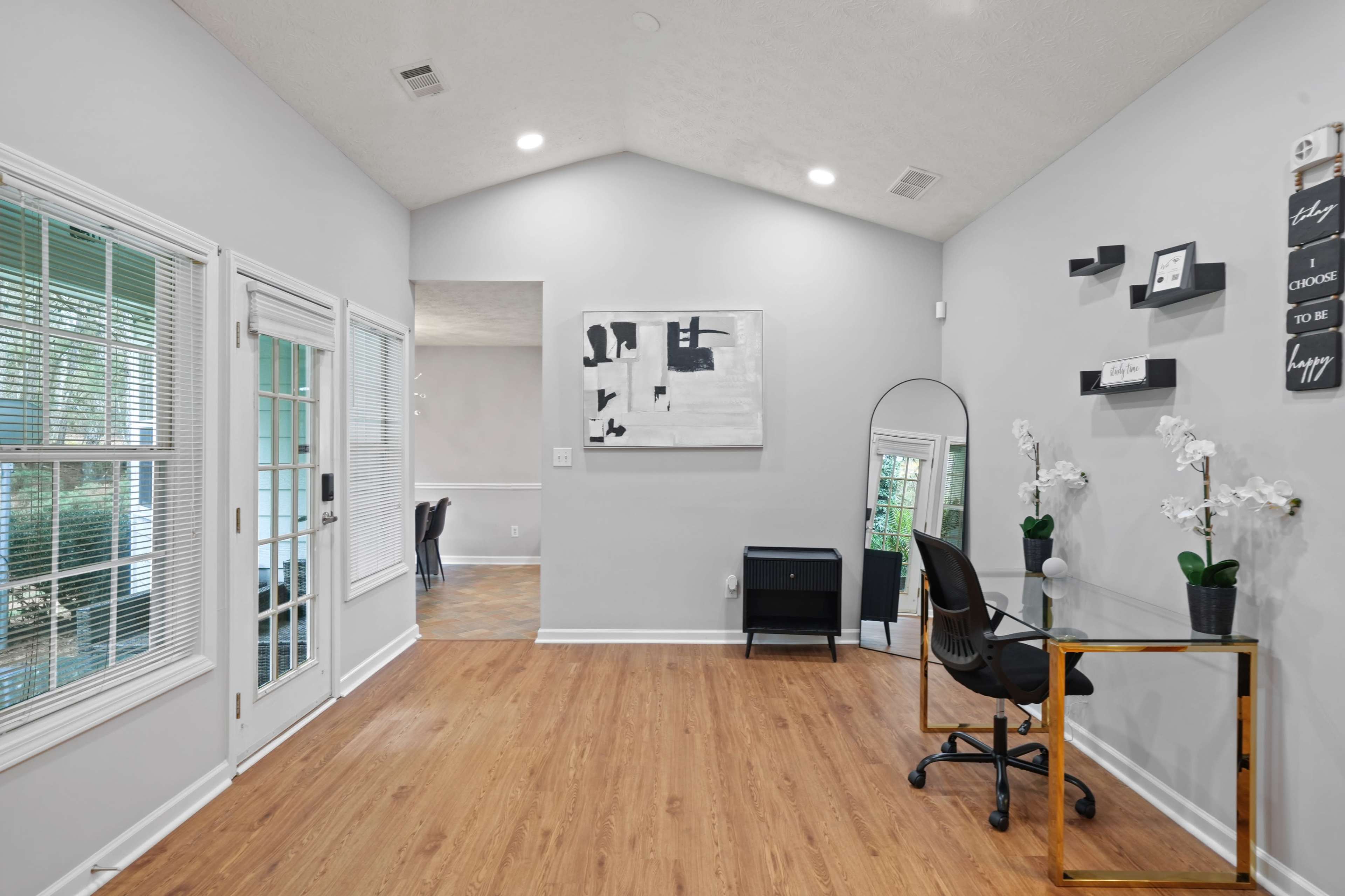The image shows a modern, well-lit room featuring a large window, a desk with a chair, and decorative elements including a mirror and wall art.