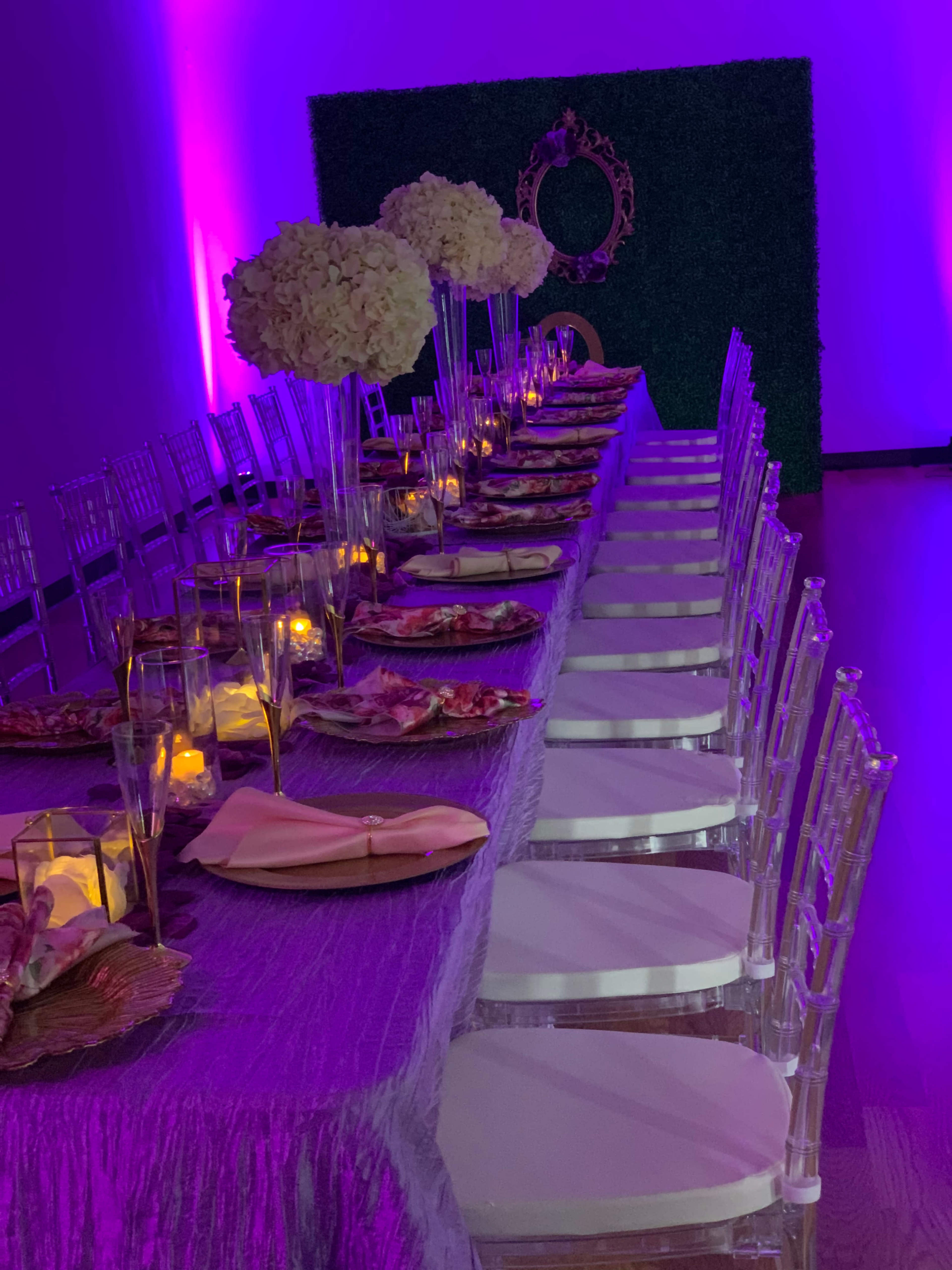 A long banquet table is set with elegant tableware, adorned with floral centerpieces and illuminated by purple lighting.