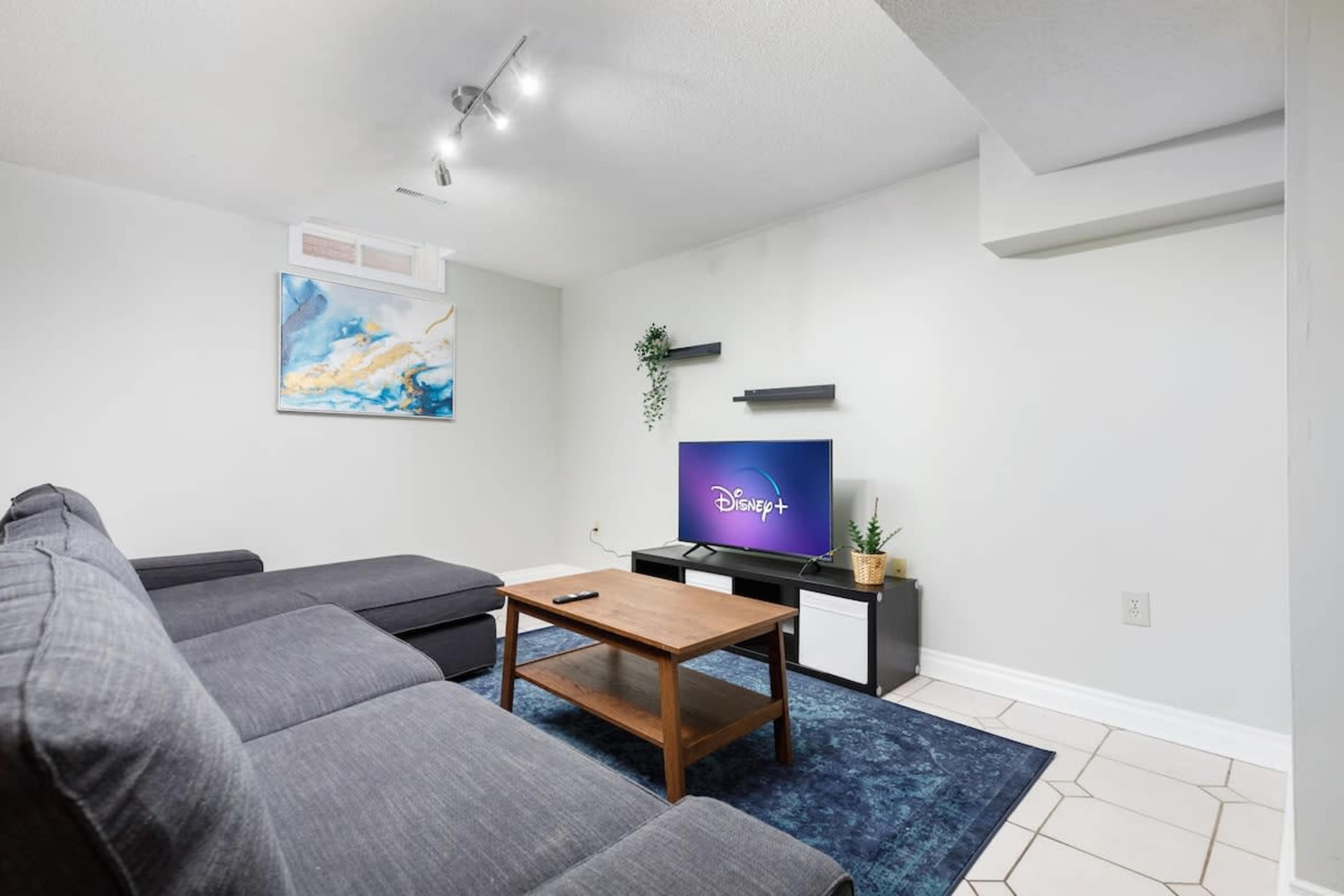A modern living room with a dark gray sectional sofa, a wooden coffee table, and a TV stand featuring a screen displaying the Disney+ logo.