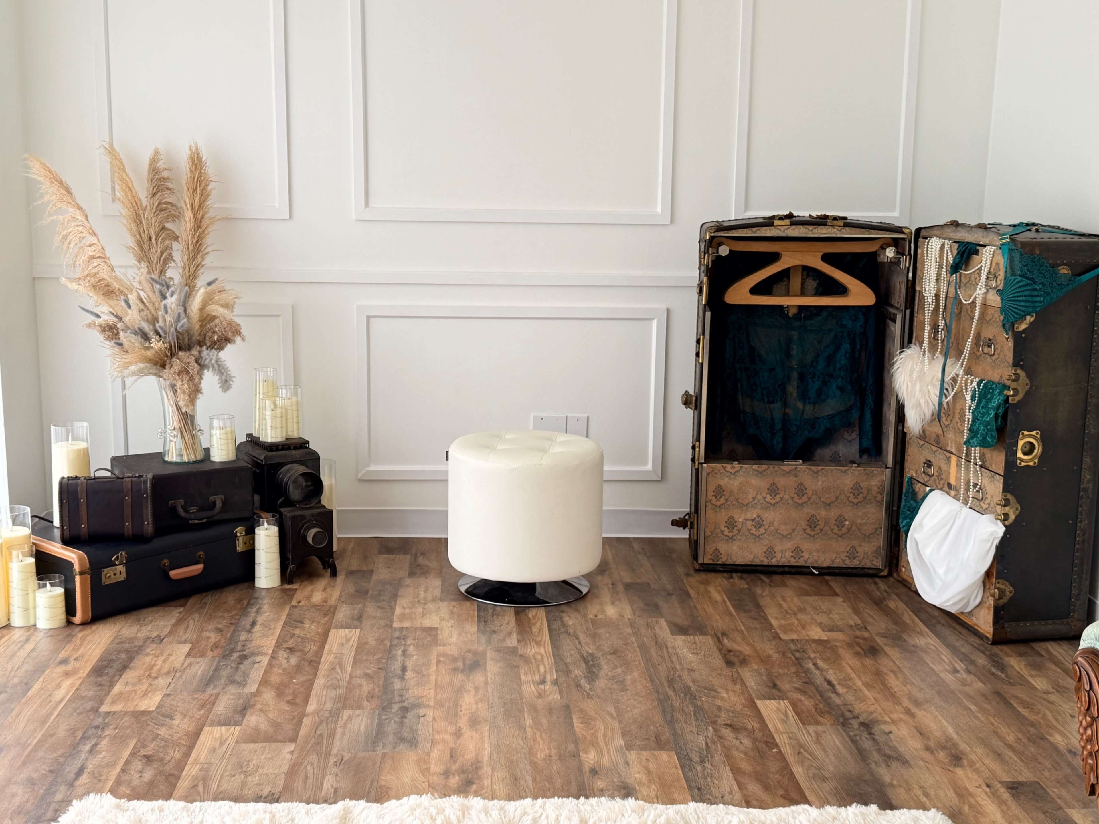 The scene features vintage suitcases and a decorative trunk alongside a modern round ottoman in a well-lit room with wooden flooring and a wall adorned with paneling.