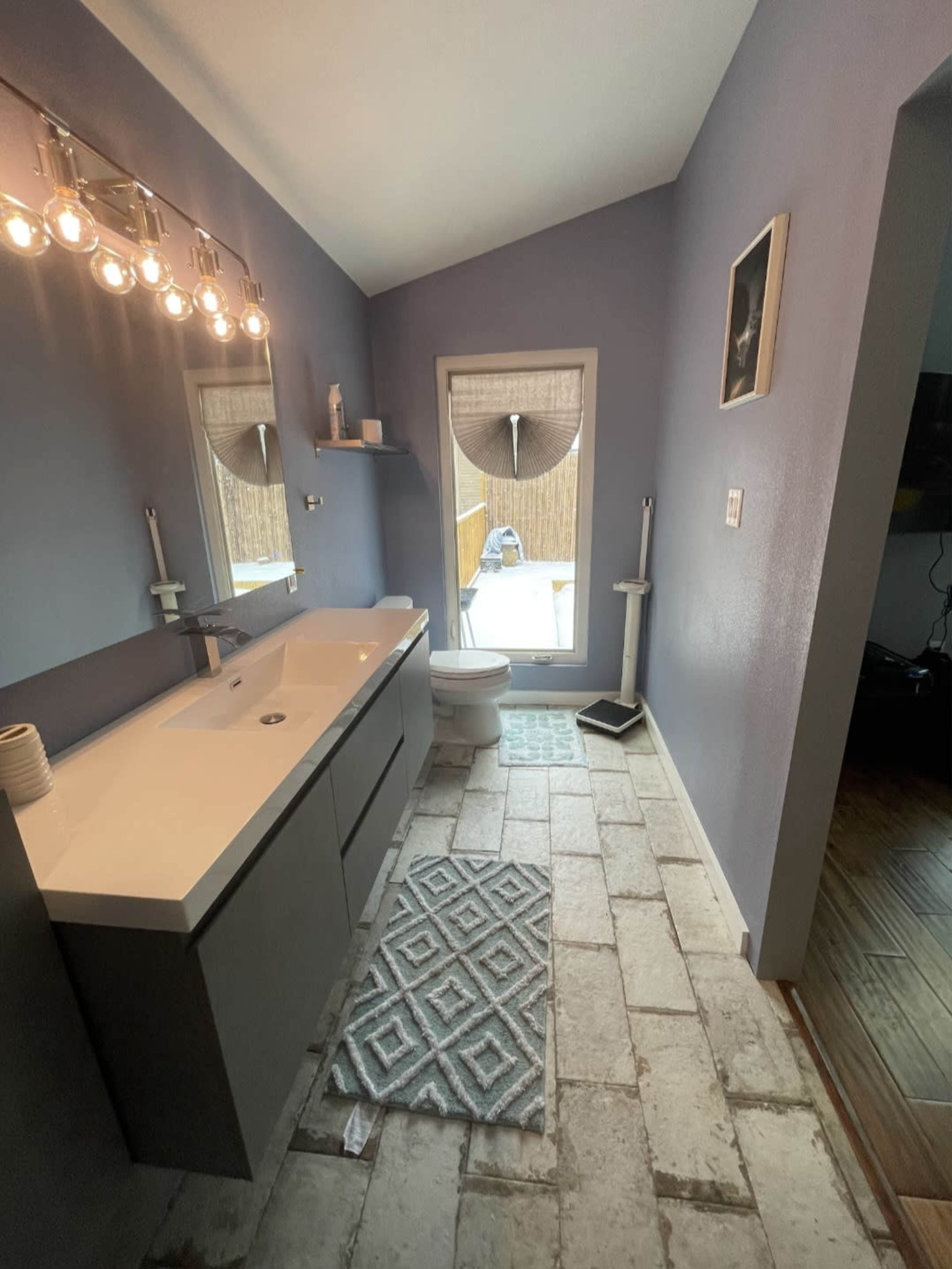 The bathroom features a modern sink vanity, a toilet, and a window with a view of an outdoor area, all set against light blue walls and tiled flooring.