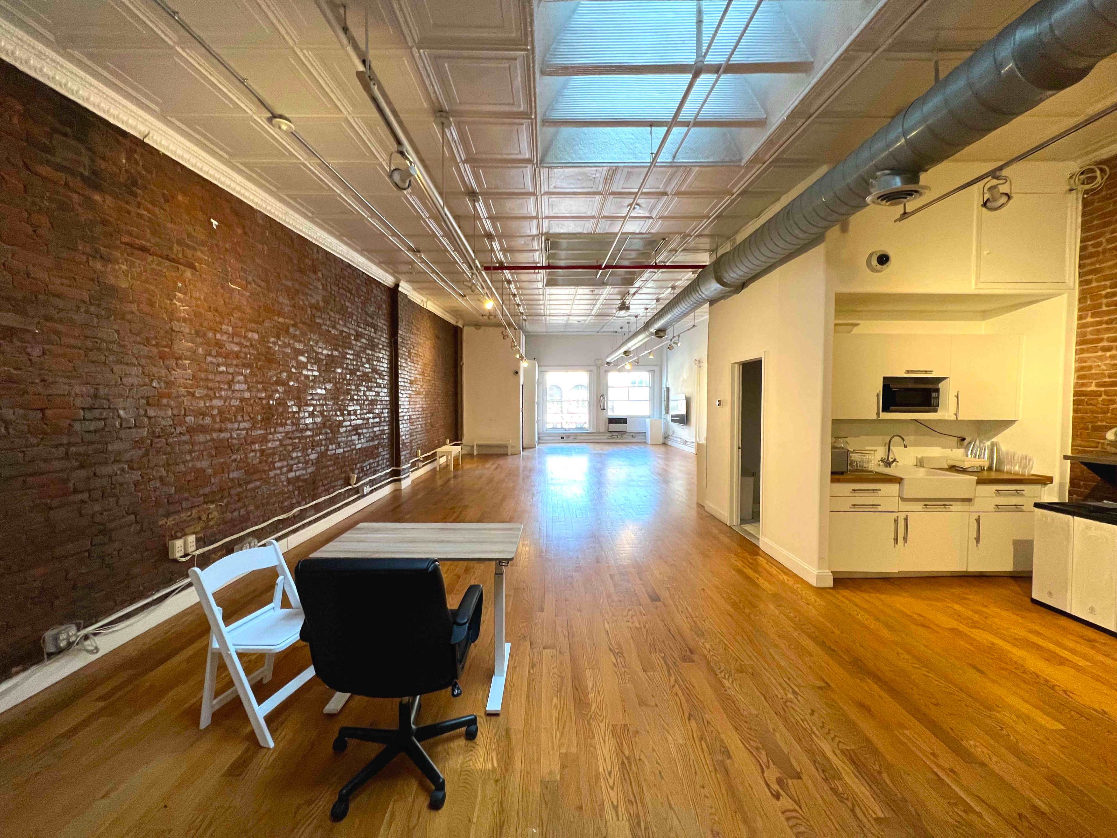 A spacious room with exposed brick walls, hardwood flooring, and a kitchen area in one corner.