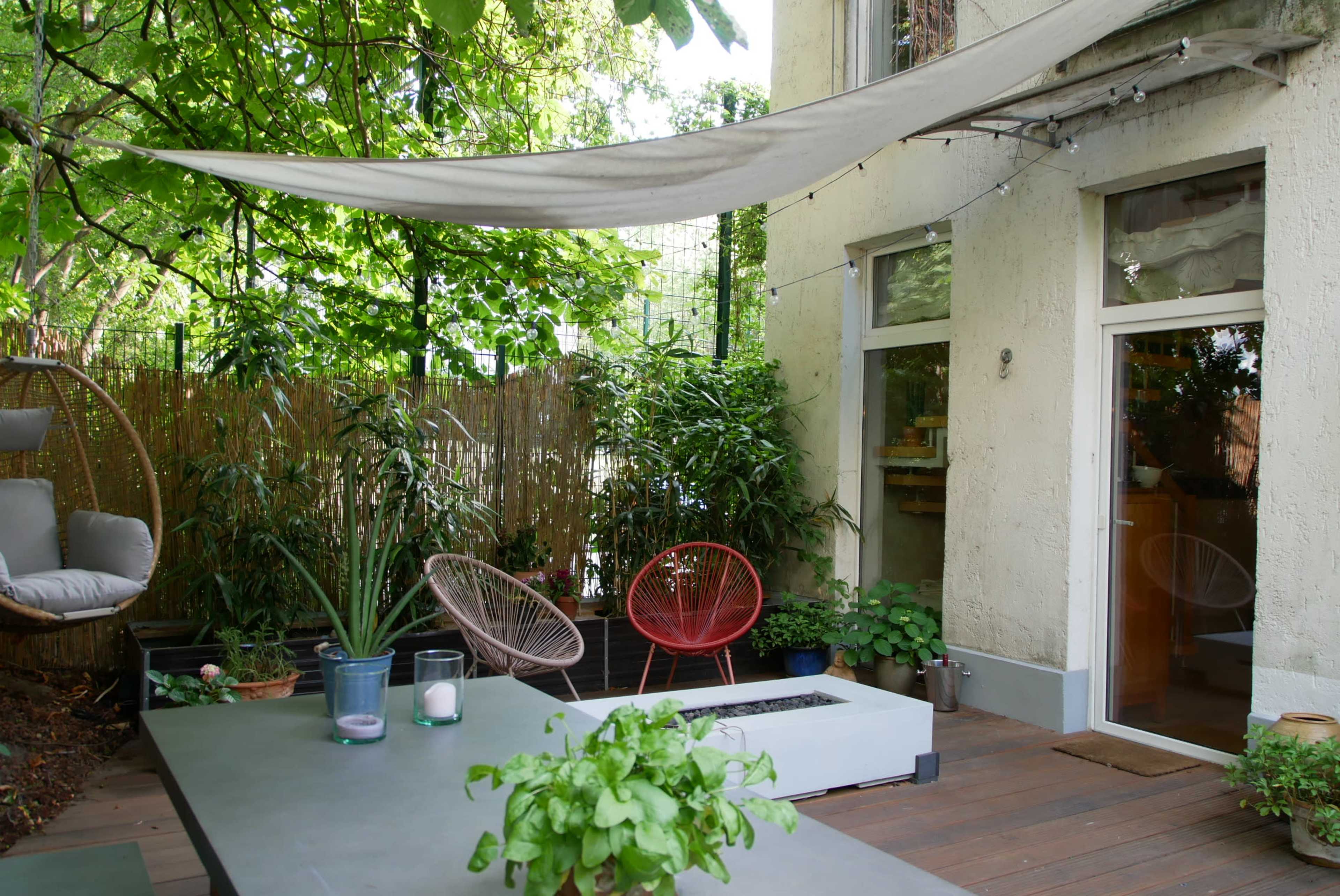 A patio area features a shaded table, potted plants, and colorful chairs surrounded by greenery and a house.