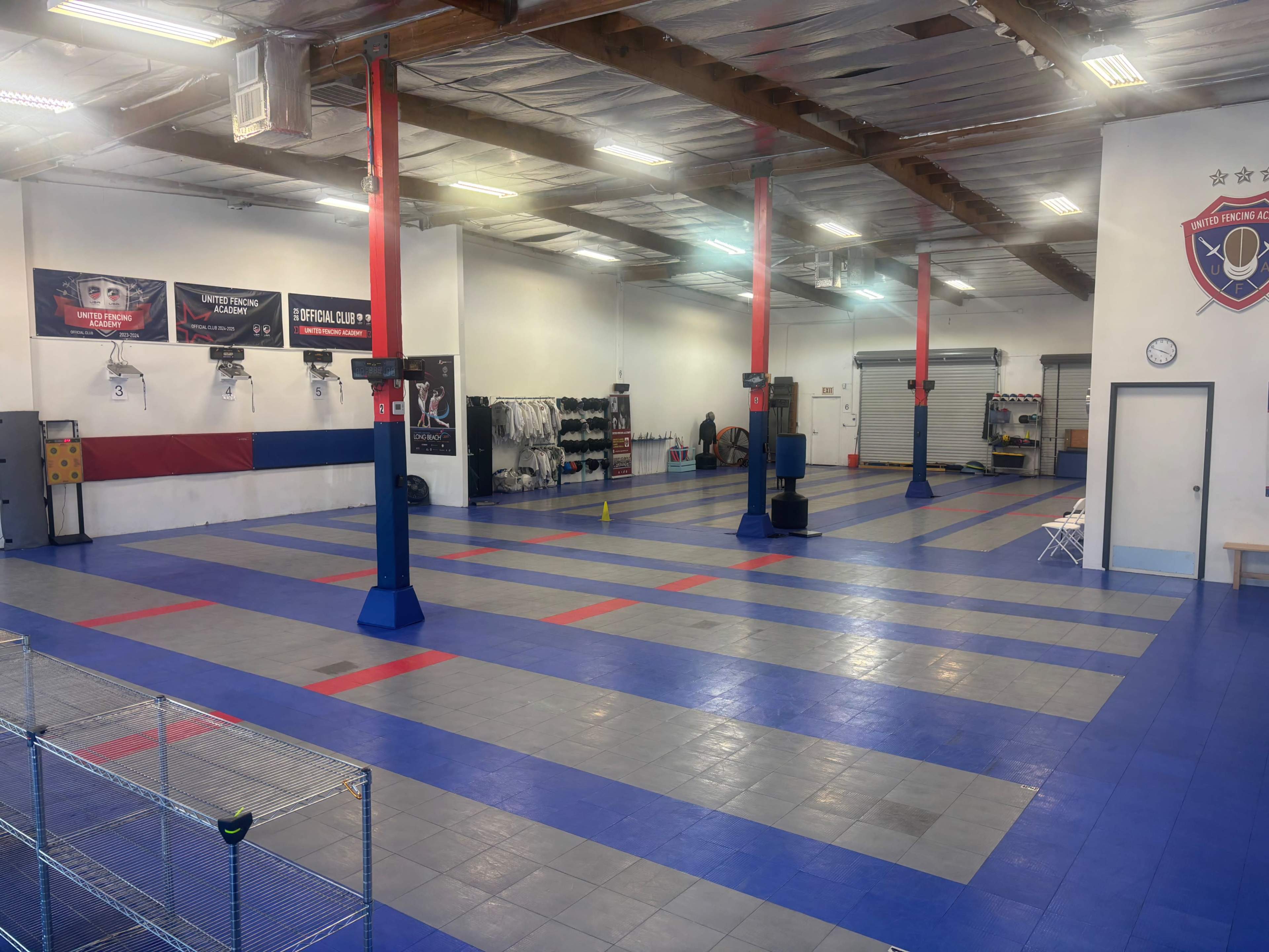 The image shows a spacious training facility with blue and gray flooring, red support pillars, and various training equipment neatly arranged along the walls.