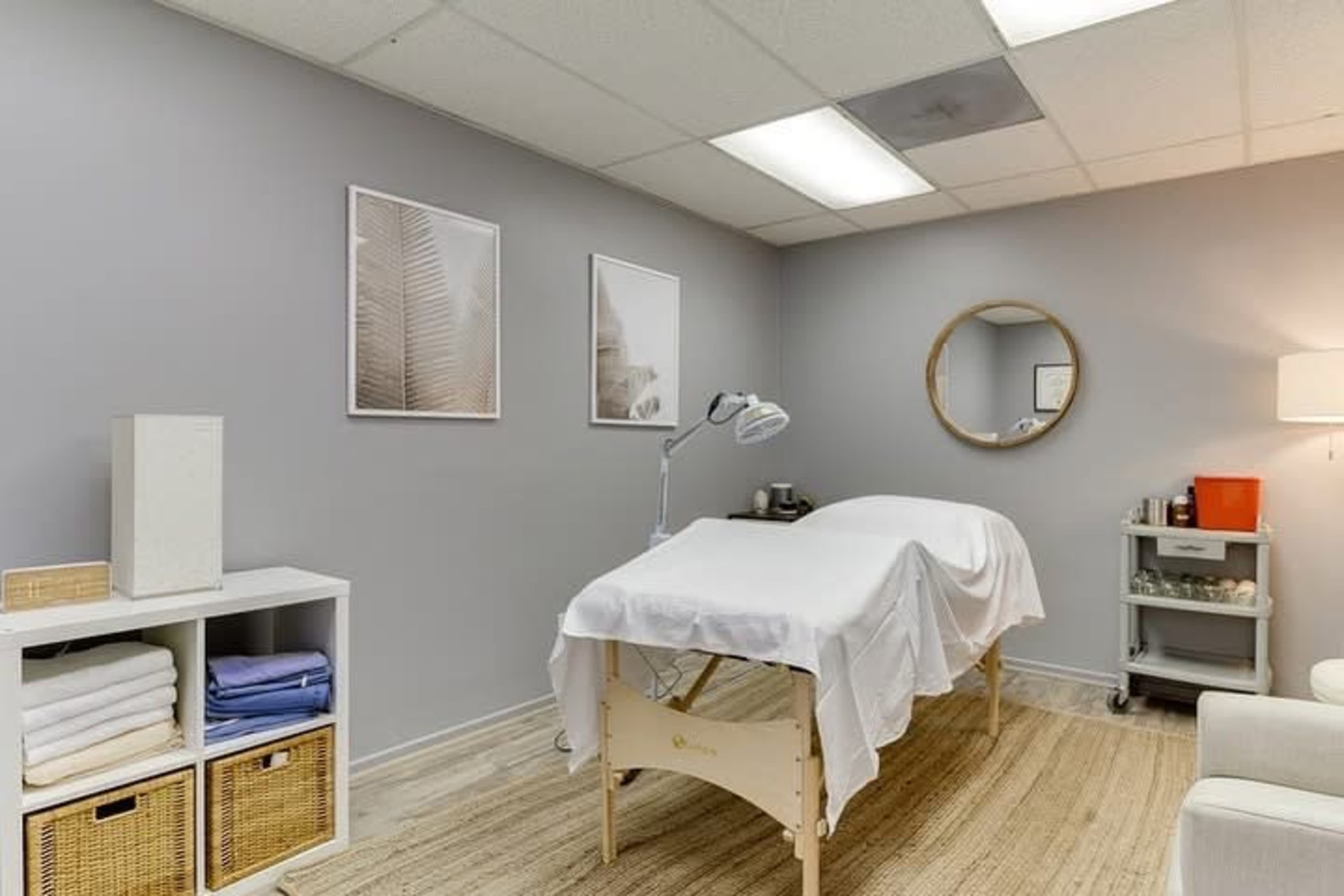 The image shows a treatment room with a massage table covered in a white sheet, surrounded by neutral-colored walls and minimal furnishings, including shelves and a lamp.