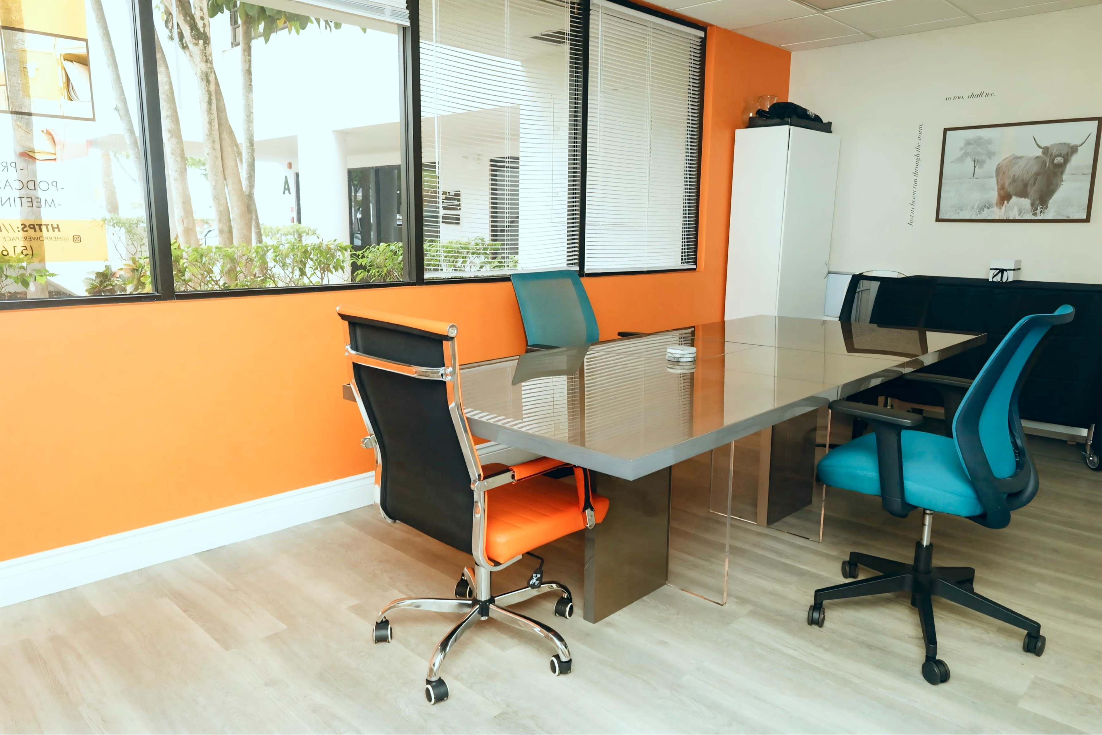 The image shows a modern conference room with a glass table and four chairs, set against an orange wall and featuring large windows that allow natural light.